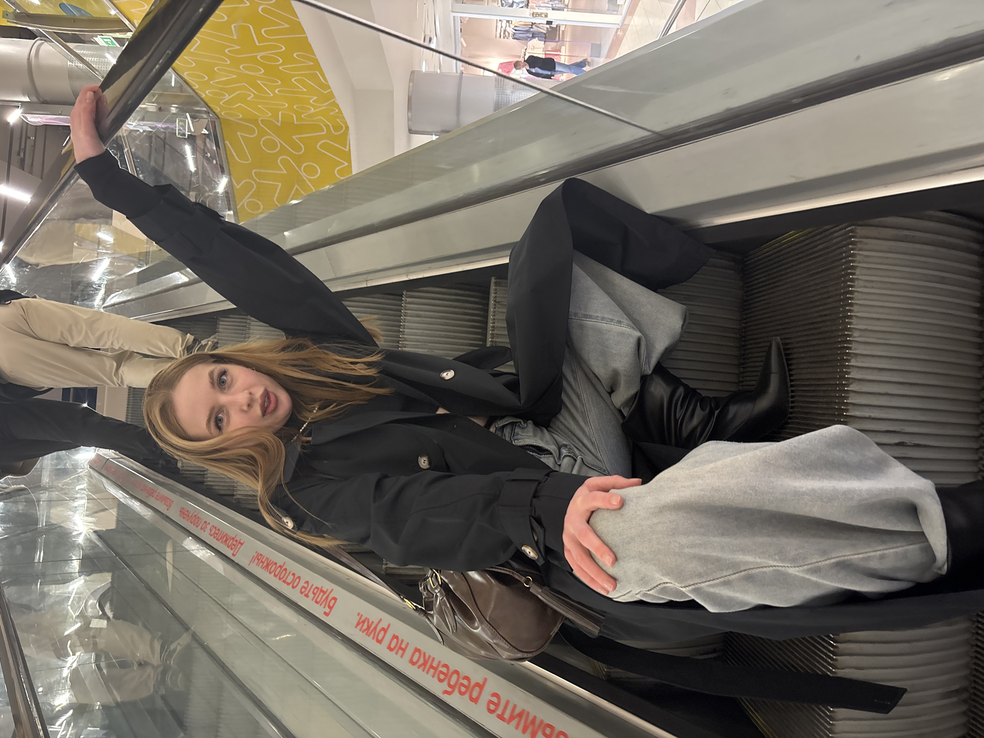 A woman lies on an escalator in what appears to be a shopping mall in Perm, Russia. She is wearing jeans, a black coat, and black boots. She is holding onto the escalator's handrail. A brown purse rests beside her on the moving steps. The escalator ascends towards a brightly lit upper level of the mall. Warning signs in Russian are visible on the escalator's sides.
