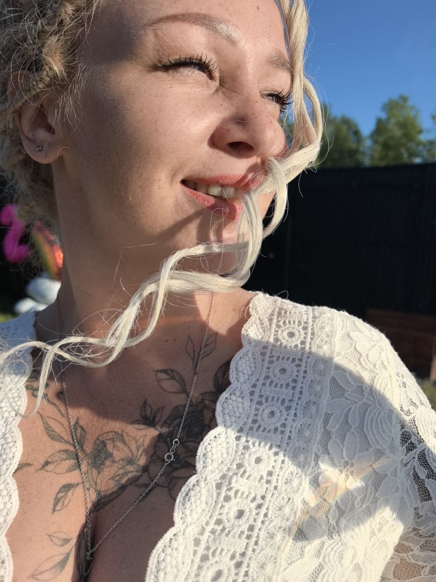 In Noviy, Russia, a woman with blonde dreadlocks is captured in a close-up shot. She is wearing a white lace top, revealing tattoos on her chest and neck, including floral designs and possibly lettering. A silver necklace with a pendant rests on her chest. Her eyes are closed, and her head is tilted upwards, with a strand of her hair blowing across her face, obscuring part of her mouth. Her lips are slightly parted, showing her teeth, and she appears to be smiling or laughing. The background is blurred, with hints of greenery and a dark structure, suggesting an outdoor setting under a clear blue sky. The sunlight casts shadows and highlights on her face and clothing.
