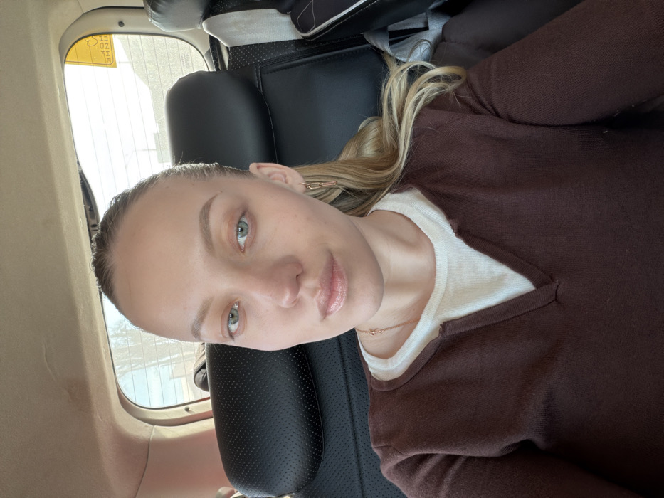 The image depicts a female individual seated in the rear passenger compartment of a vehicle, facing the camera. Her head is centered, and her gaze is directed forward. She has light skin, light-colored eyes, and her light-brown hair is pulled back, with some strands visible over her right shoulder. She is wearing a dark brown long-sleeved V-neck top over a light-colored V-neck undershirt. A delicate gold-colored chain necklace with a small pendant is visible on her neck, and a gold-colored elongated hoop earring is present in her right ear. The interior of the vehicle shows black headrests and a light-colored ceiling. Through the rear window, an indistinct outdoor environment with bright sky is visible. A yellow rectangular sticker with black Cyrillic text, consistent with "РЕБЕНОК" (Child), is affixed to the upper right corner of the rear window. The specific location within the city of Zavidovo, Russia, cannot be confirmed from the visual evidence.