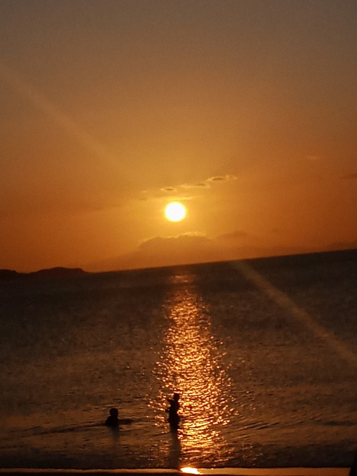 A vibrant sunset dominates the sky, casting an orange glow over the sea. The sun, a bright orb, sits above low-lying clouds. Its reflection creates a shimmering path on the water's surface, leading towards the foreground where two figures stand. One appears to be wading, the other partially submerged. A dark landmass is vaguely visible on the left horizon.