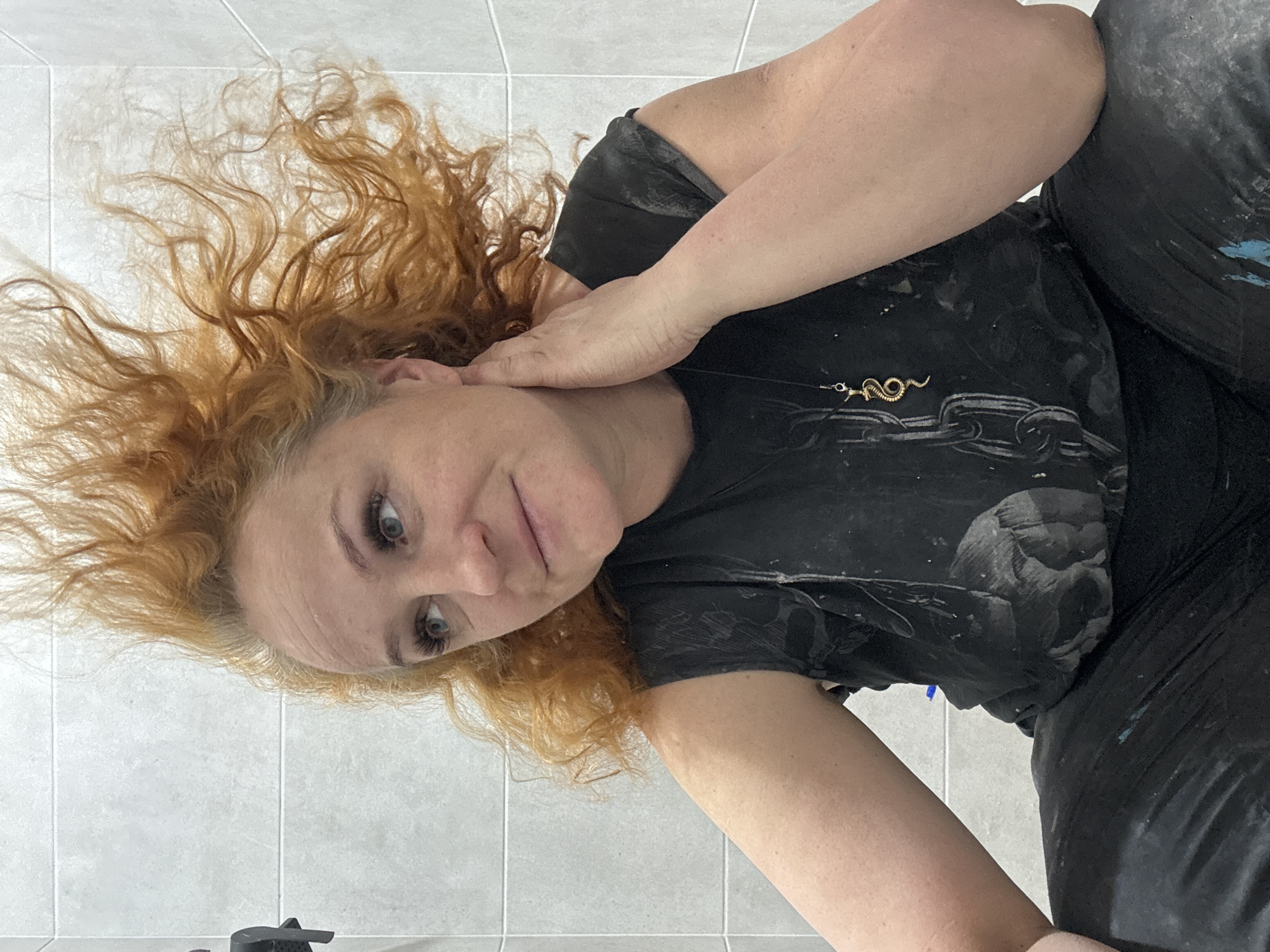 The image shows a woman with curly red hair. She is wearing a black t-shirt with a print and black pants. Her right hand is near her face. A gold snake necklace is visible on her chest. The background is a white tiled wall. The picture appears to have been taken from a low angle. There is no evidence the photo was taken in the city of Chaikovsky, Russia.