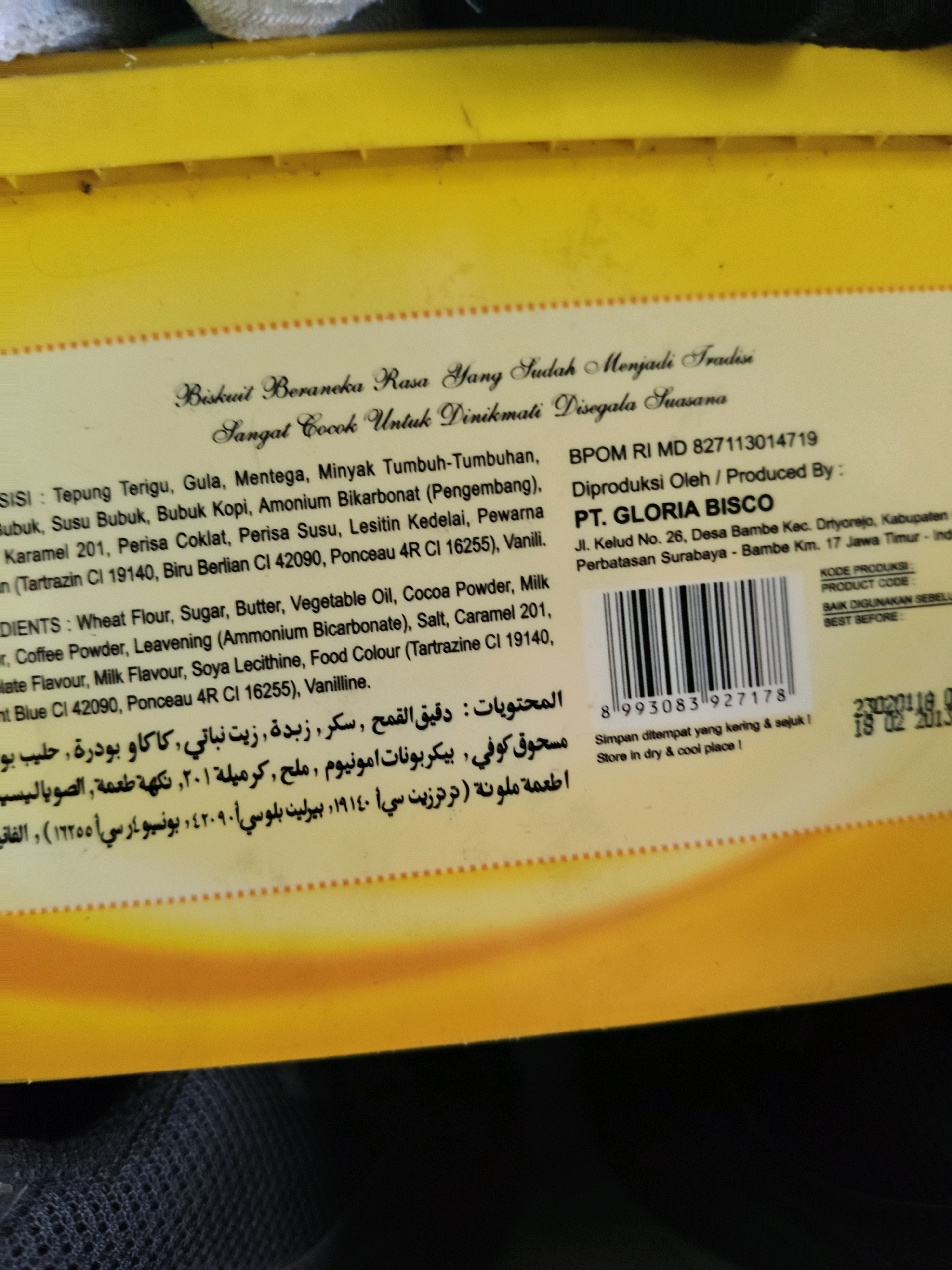 The image is a close-up of the packaging of "BISKUIT BERANEKA RASA" (Assorted Flavor Biscuits) from PT. GLORIA BISCO. The packaging is predominantly yellow and features text in Indonesian, English, and Arabic script. The Indonesian text describes the biscuits as being suitable for all occasions and lists ingredients such as wheat flour, sugar, butter, vegetable oil, cocoa powder, and various flavorings. The English text reiterates the ingredients and provides production details, including the company name and address: Jl. Kelud No. 26, Desa Bambe Kec. Dryorejo, Kabupaten Gresik, Jawa Timur, Indonesia. The Arabic script appears to be a translation of the ingredients or product description. A barcode is present on the packaging, along with a "BEST BEFORE" date of 02 2013. The location mentioned in the text, Kabupaten Gresik, Jawa Timur, Indonesia, places the product's origin in East Java, Indonesia. The image itself is taken from an angle that shows the top and side of the product packaging, which appears to be a box or container. The background is blurred and out of focus, with a dark, textured surface visible at the bottom. There are no people or any environmental context outside of the product packaging itself visible in the image. The context of "Sumberwono, Indonesia" is not directly evident from the image content, other than it being part of the broader geographical information of East Java, Indonesia, where the product is manufactured.