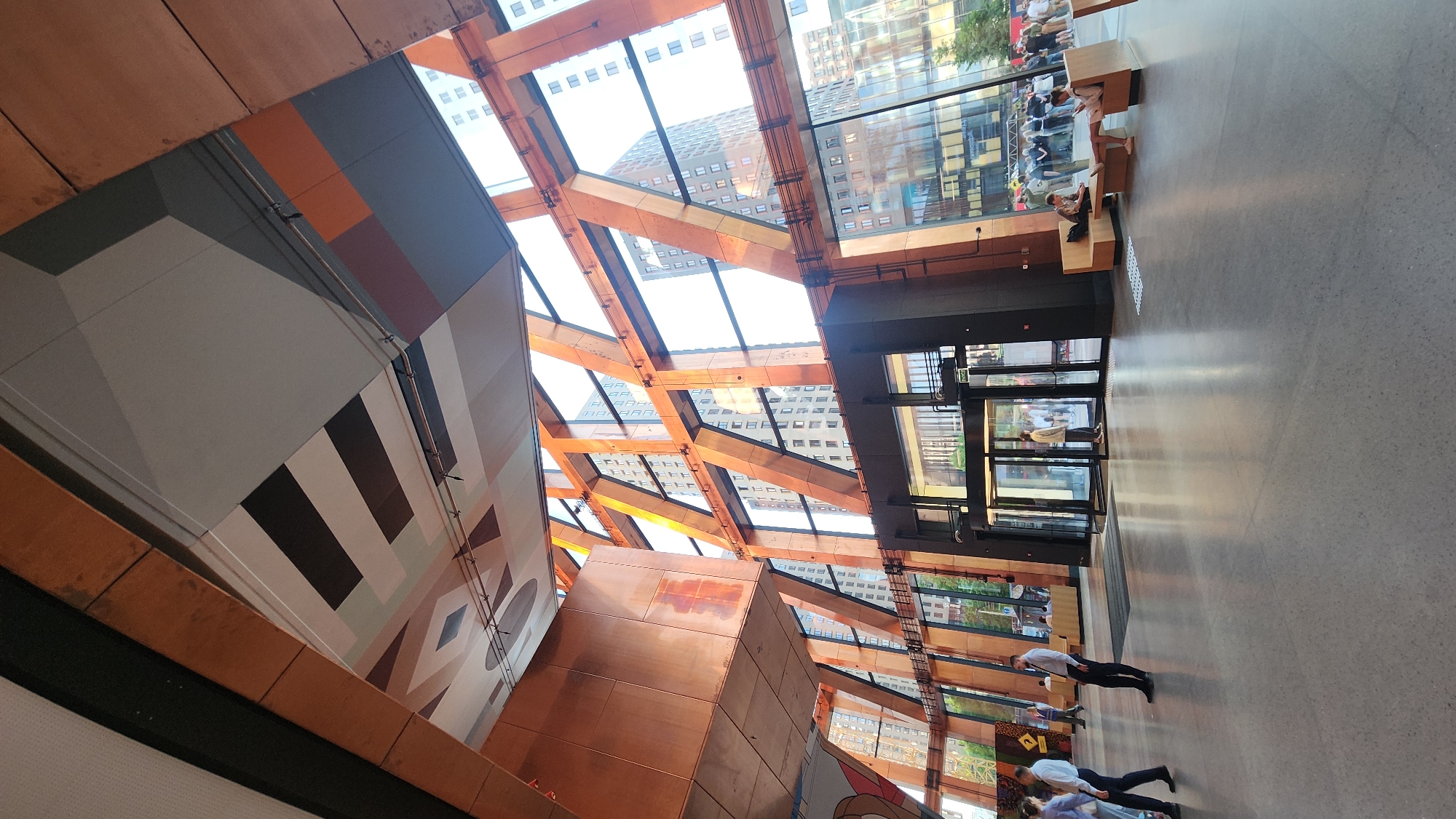 The image shows an interior view of a building in Moscow, Russia. The building features a modern architectural design with a glass ceiling supported by a wooden frame. Buildings are visible through the glass roof, indicating an urban location.
The interior includes a decorated wall with geometric shapes and colors. A metallic structure is also visible near the wall.
The floor appears to be polished concrete. There are people walking toward and entering through glass doors. Seating is present along the windows on the right side of the image.