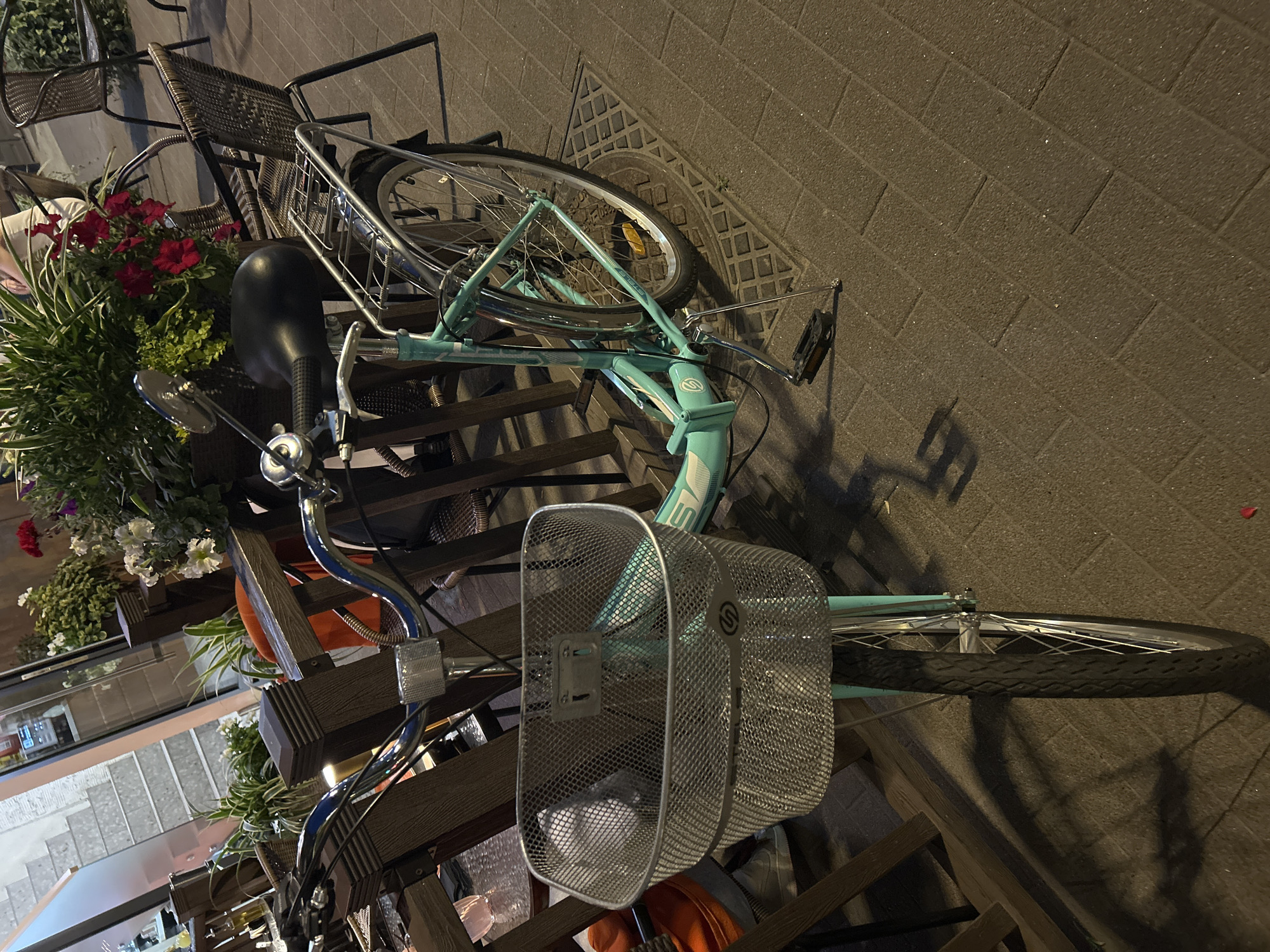A teal bicycle with a silver basket on the front and a silver rack on the back is parked on a brick sidewalk. The bicycle is facing left and is slightly tilted. The handlebars are silver and the seat is black. There are plants in the background, and a table and chairs are also visible. The scene is outdoors, and it appears to be evening. The location is Unknown, Unknown.