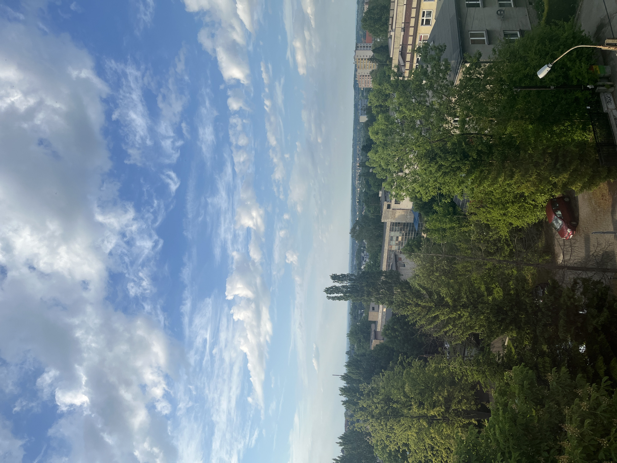 The image depicts an outdoor urban scene under a sky filled with white cumulus clouds. On the right side of the image, a partial view of buildings is visible, including a high-rise structure and smaller adjacent buildings. A dense cluster of green trees partially obscures some of the building facades. A lamppost is positioned on the right, and a red car is parked on the roadside.