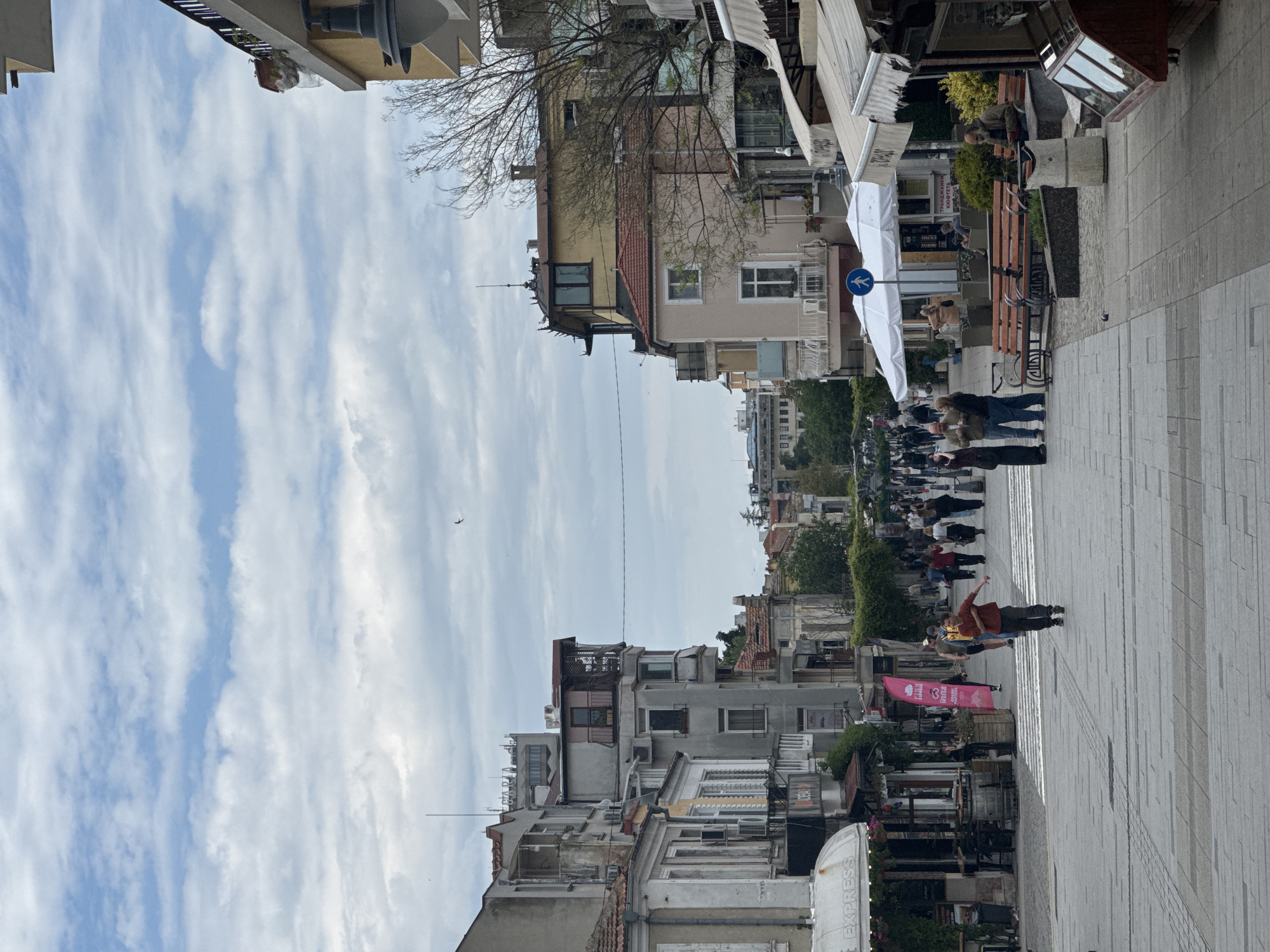 The image depicts a bustling pedestrian street in Burgas, Bulgaria. The view is from ground level looking toward the horizon. Buildings line both sides of the paved street. The sky, predominantly blue with streaks of white clouds, occupies the top portion of the frame.
On the right side of the street are benches and people. Several individuals and small groups are walking along the street. A couple appears to be dancing or gesturing in the foreground on the right side of the street.
On the left side, buildings of varying heights and architectural styles line the street. Some buildings show signs of aging or disrepair. A pink sign with writing is visible on the lower-left side, as are outdoor cafe umbrellas and structures.
The scene suggests a lively urban environment with a mix of commercial and residential structures.