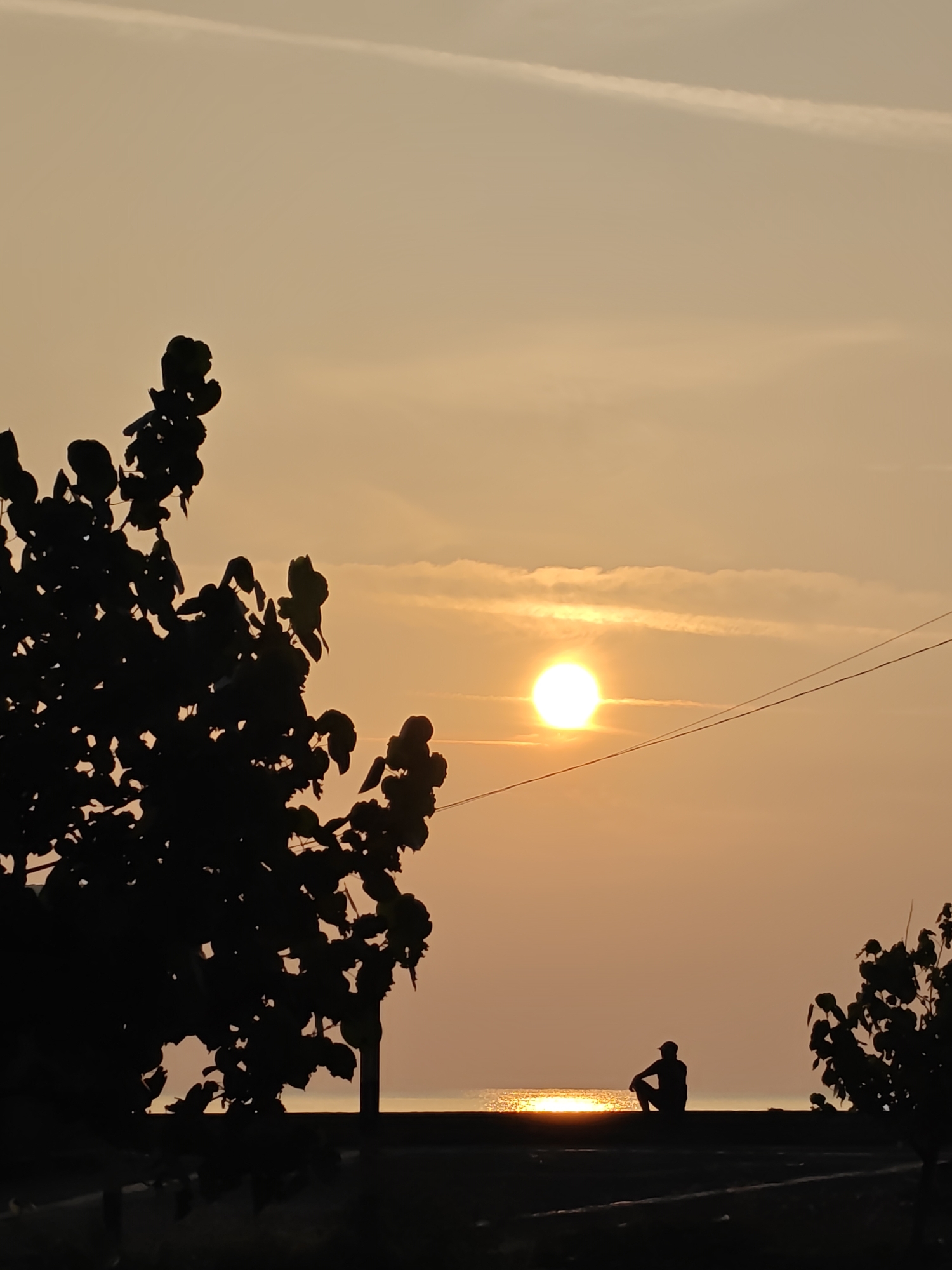 This photograph captures a sunset over the ocean, as viewed from a coastal area in Lhokseumawe, Indonesia. In the foreground, the dark silhouettes of trees frame the scene on the left and right. A single person, also silhouetted, is seated on a raised structure facing the water, with their back towards the viewer. The sun, a bright orb, is positioned in the middle of the frame, just above the horizon, casting a shimmering reflection on the water's surface. Thin clouds are visible in the sky, and a power line stretches diagonally across the upper right portion of the image. The overall atmosphere is tranquil and serene, characterized by the warm hues of the sunset.