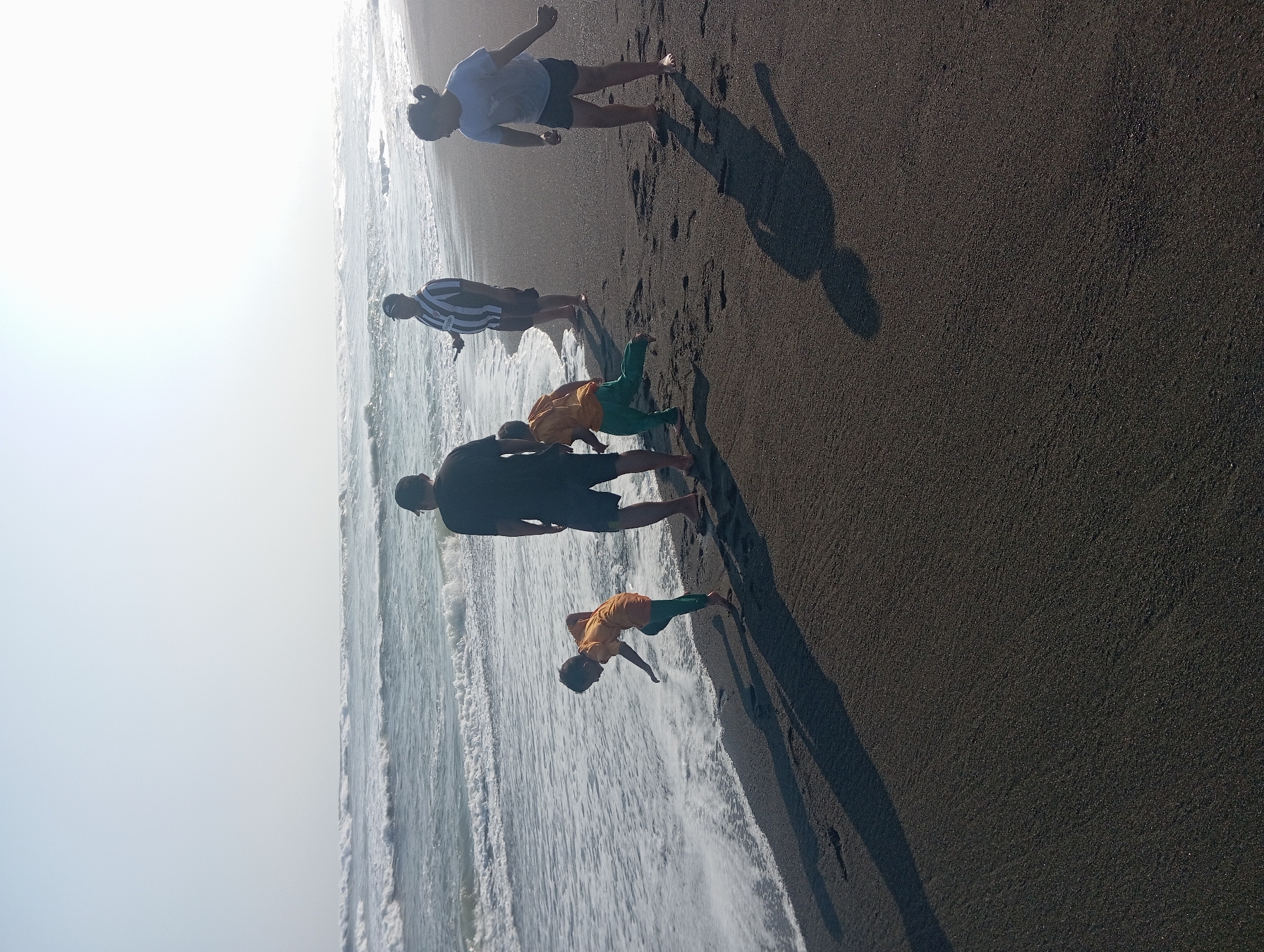 This image depicts a group of people on the black sand beach of Parangtritis, Indonesia, with the ocean and waves in the background. There are at least five individuals visible. In the foreground, a child wearing an orange shirt and green pants is splashing in the shallow water at the edge of the sea. To the right of this child, another child, possibly a girl with dark hair pulled back, is wearing a light-colored t-shirt and dark shorts and appears to be watching the waves. Further back, a man in a black t-shirt and shorts stands near the water. To his left, another man is wearing a black and white striped shirt and shorts. A fifth person, a child wearing an orange shirt and green pants, is running towards the water, with their back to the camera. The sun appears to be low in the sky, casting long shadows on the dark sand. The waves are breaking on the shore, creating white foam. The sky is bright and hazy, indicating either early morning or late afternoon light.