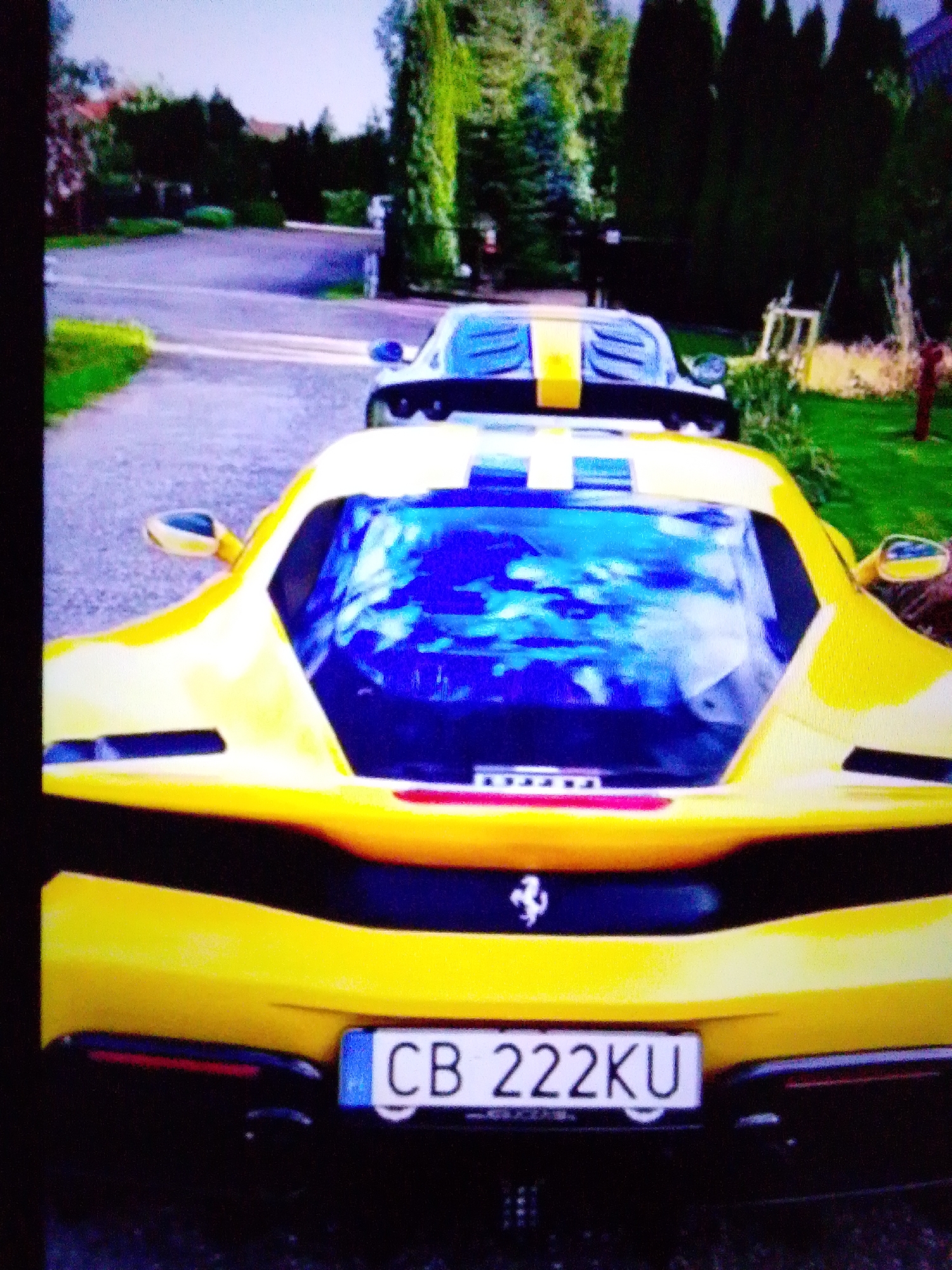 A yellow Ferrari is parked in front of a green lawn and trees. A second car, also yellow with a black and yellow stripe, is visible in the background. The license plate on the yellow Ferrari reads "CB 222KU". The image is taken from a low angle, looking up at the car. The lighting suggests it is daytime, with sunlight reflecting off the car's surface. The environment appears to be suburban, with houses and trees visible in the distance. The exact location is unknown but it is a private residence.