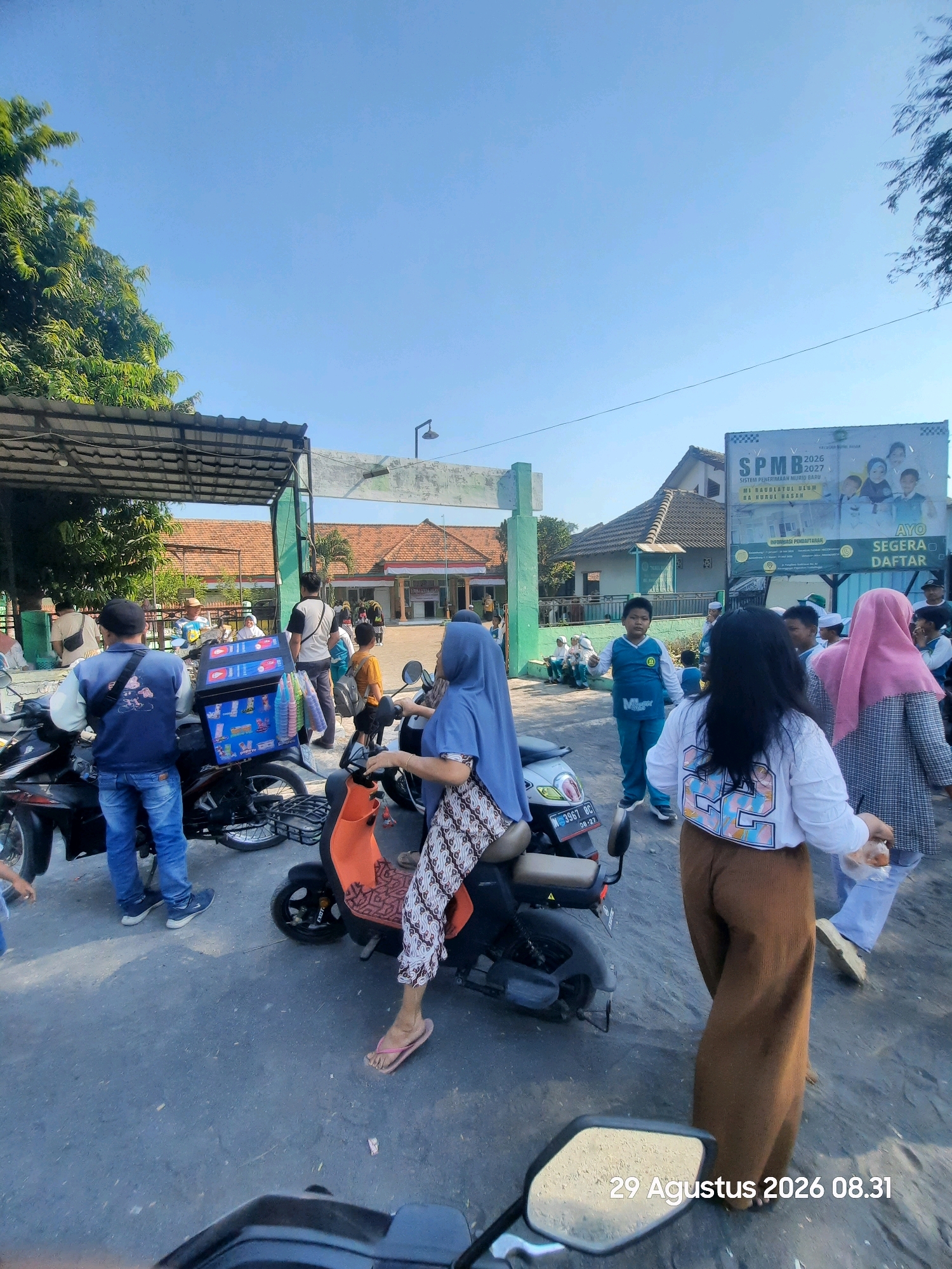 In an outdoor scene in Sukokerto, Indonesia, on August 29, 2026, at 08:31, a group of people are gathered near a building entrance. The sky is clear and blue. In the foreground, a person on a scooter, wearing a blue headscarf and a patterned dress, is looking forward. To their left, a person in a blue jacket and jeans is standing by a parked motorcycle. A blue box with images of ice cream is attached to the back of a scooter. Further back, several individuals, including children wearing uniforms, are standing and walking. A large banner on a building in the background displays text in Indonesian, including "SPMB 2026-2027 SISTEM PENERIMAAN MURID BARU" and "AYO SEGERA DAFTAR." The environment appears to be a school or educational institution, with buildings featuring red-tiled roofs and paved ground. Some trees are visible on the left side of the image.