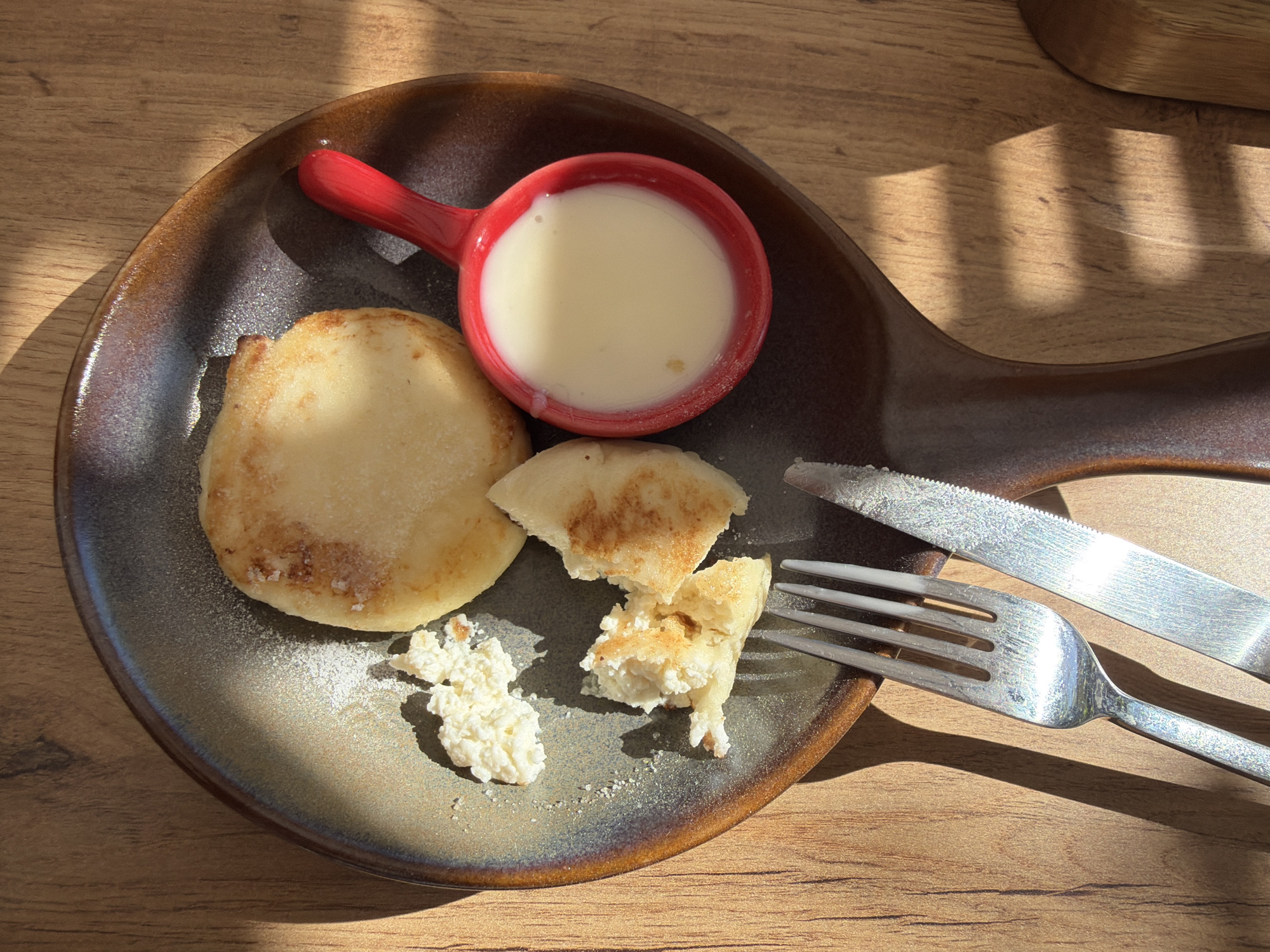 A top-down shot shows a plate with food on a wooden table, likely taken indoors. The plate, a dark brown color with a slight bluish-grey tint, contains what appears to be a pancake or cheese pancake and some smaller pieces of the same. Next to the pancake is a small red handled bowl containing a white liquid, likely sour cream or condensed milk. A fork and knife lie on the plate beside the food. The table is a light brown, possibly made of wood. Sunlight is shining in, casting shadows on the table and the plate. The location is Chaikovsky, Russia.