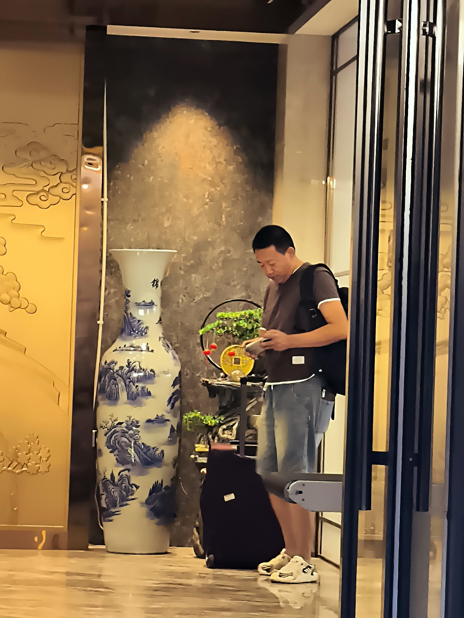 A man stands indoors, looking at his phone. He has short dark hair and is wearing a dark t-shirt, denim shorts, and white sneakers. A black backpack is slung over his right shoulder. To his left is a tall, ornate vase with a blue and white landscape design. A small bonsai tree with red ornaments sits on a shelf to the right of the vase, near the man. A dark suitcase is on the floor next to the man. The environment appears to be a hotel lobby or a similar indoor public space, with warm lighting and decorative elements. The image does not provide specific information about the city of Ürümqi, China, or its location within the city.