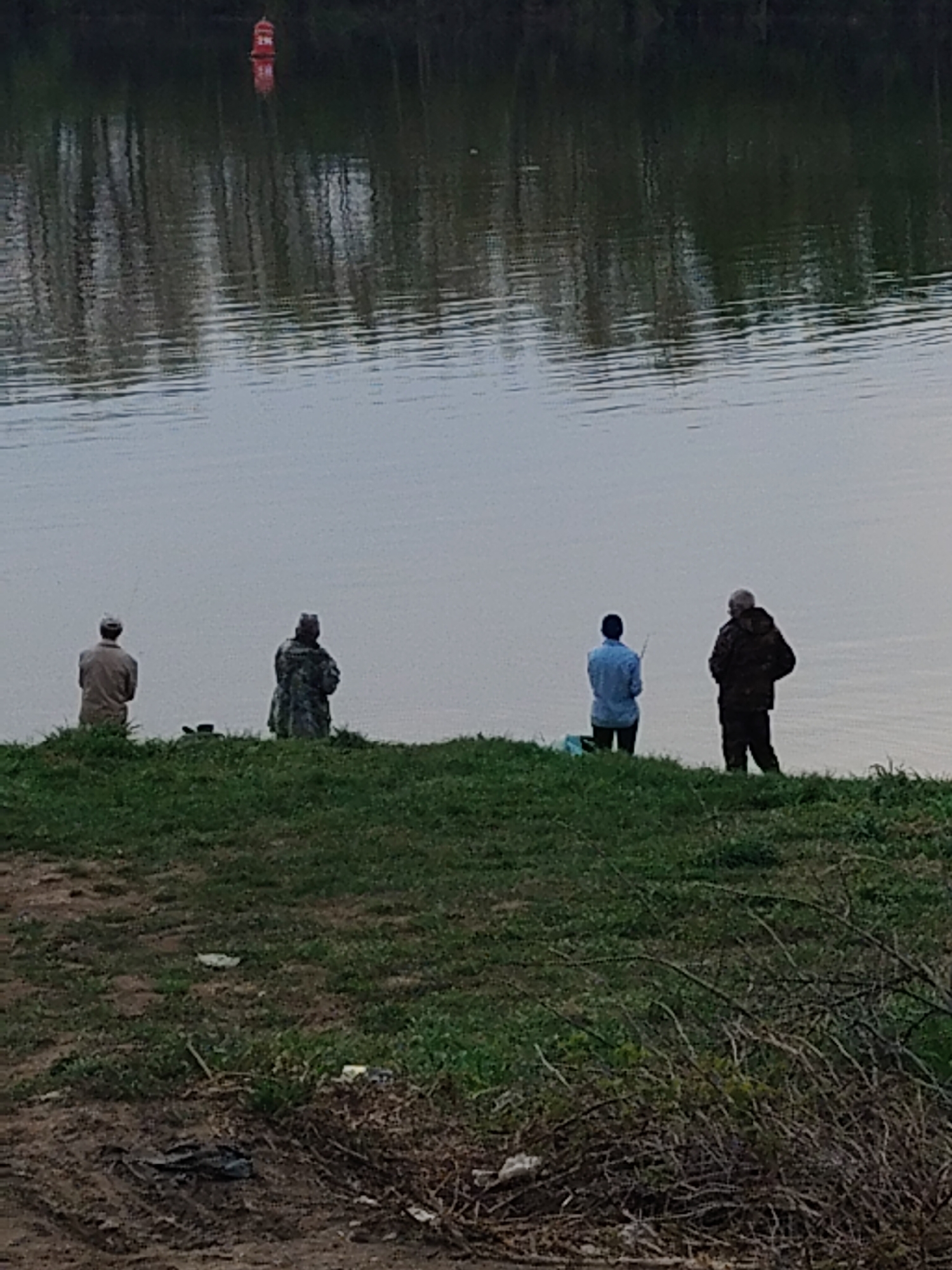 Four individuals are fishing along a body of water. The water reflects trees along the opposite bank, and a red navigational buoy is visible. The fishermen are standing on a grassy bank. The location is Unknown, Unknown.