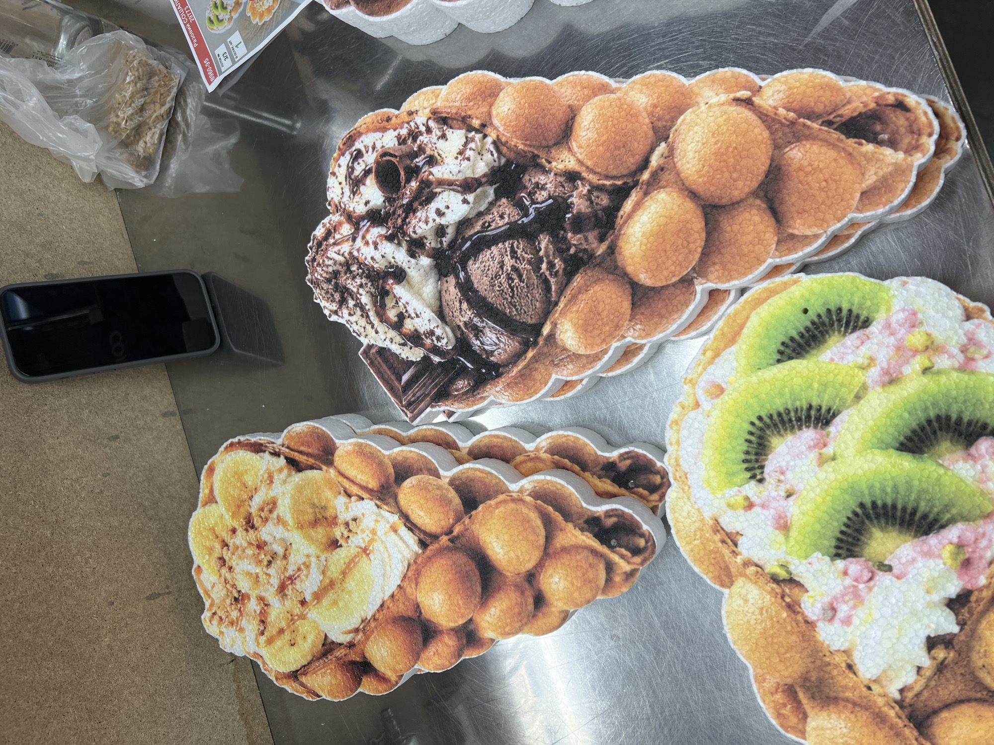 The image shows three large, cutout advertisements for bubble waffles, each topped with different ingredients. The advertisements are stacked on top of each other on a metallic surface. The top advertisement features a bubble waffle filled with whipped cream and topped with sliced bananas and what appears to be chopped nuts and caramel sauce. The middle advertisement displays a bubble waffle filled with chocolate ice cream, whipped cream drizzled with chocolate sauce, and a piece of chocolate. The bottom advertisement showcases a bubble waffle with whipped cream, sliced kiwi fruit, and possibly some pink sprinkles or edible flowers. In the background, a dark-colored smartphone is leaning against a wooden surface, and a plastic bag filled with granola or cereal is visible. A price list or menu is also partially visible in the upper right corner. The overall scene suggests a retail environment, likely a food establishment.