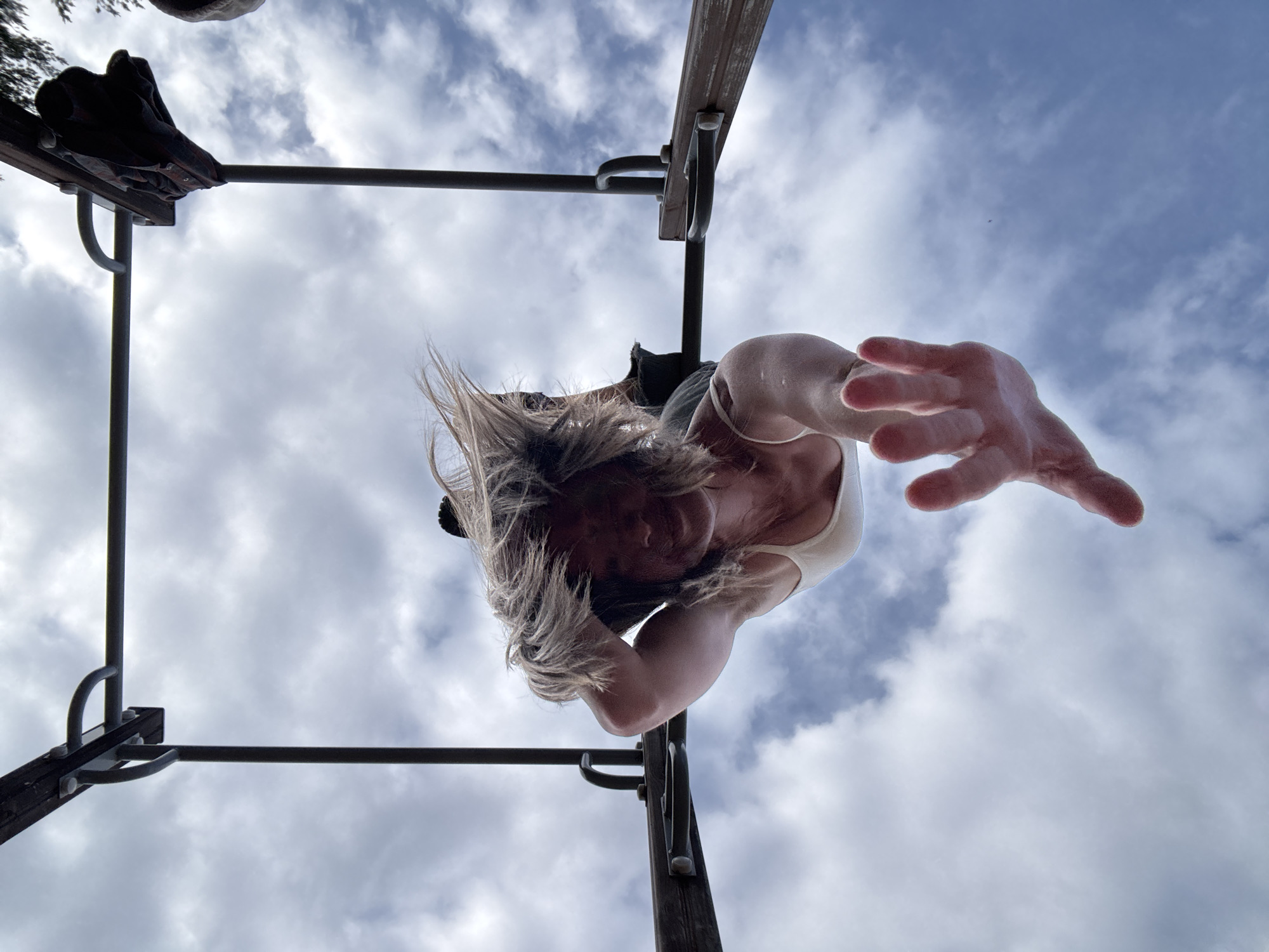 The image is taken from a low angle, looking upwards towards a person hanging upside down from a metal and wood structure, likely a jungle gym or exercise bars. The person is Caucasian, female, with blonde hair, wearing a white tank top. Her right hand is extended towards the camera. A piece of dark clothing or gear is visible on the bars above her. The background is a cloudy sky. The location appears to be outdoors. Specific coordinates and location in the city of Chaikovsky, Russia are unknown from the image.