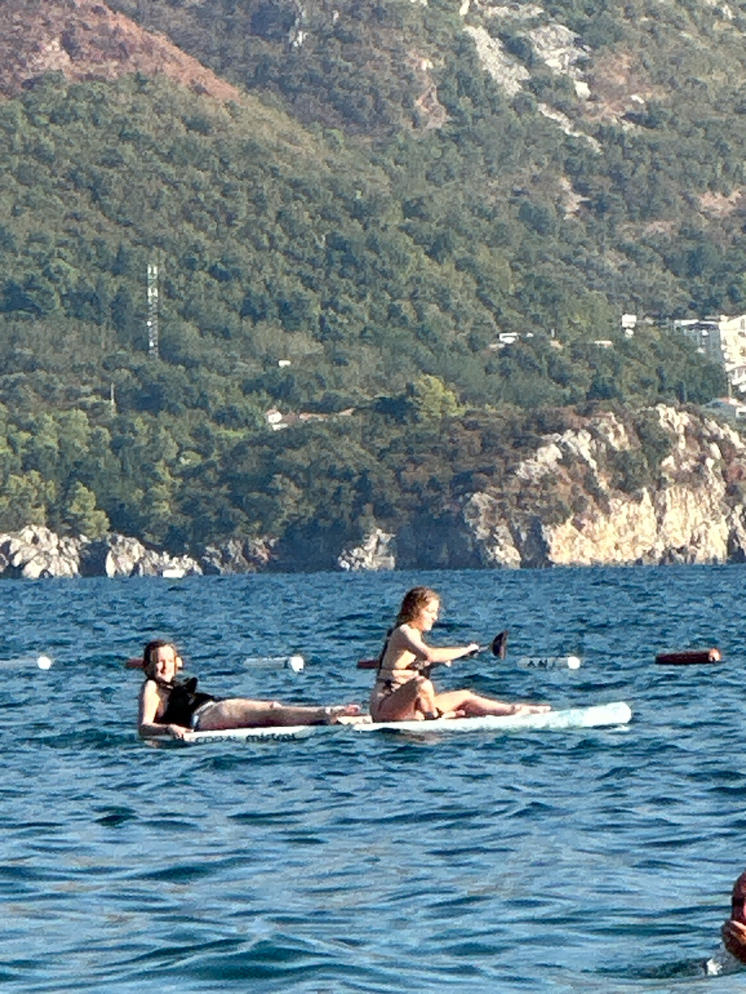 Two individuals are engaged in paddleboarding on the Adriatic Sea near the city of Boreti, Montenegro. The first individual, positioned on the left, is lying down on a paddleboard, facing the camera. They are wearing a dark swimsuit. The second individual, on the right, is seated on another paddleboard, holding a paddle and facing left. They are wearing a bikini. Both paddleboards are white and appear to be floating on calm, blue water. In the background, a verdant, mountainous coastline with scattered buildings is visible. The terrain is characterized by dense foliage and rocky outcrops. The overall environment suggests a sunny day at the seaside.