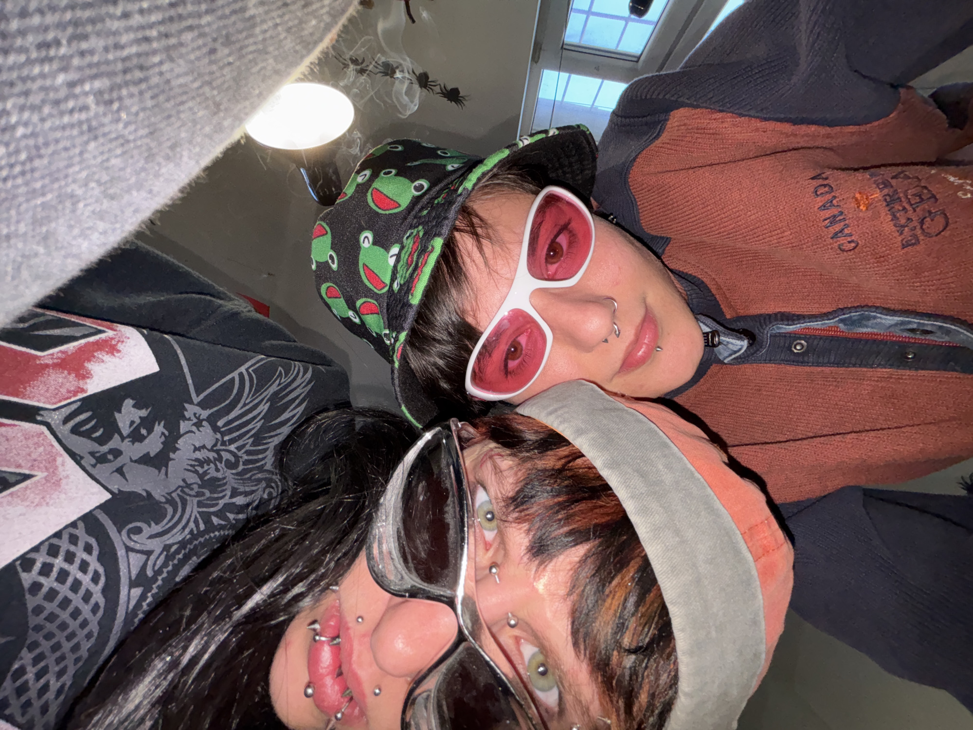 Two people are in an indoor selfie. One person wears a black bucket hat with green frogs, white-framed red sunglasses, and a brown and blue sweater with "CANADA" stitched on the chest. The other person wears black sunglasses, a gray and orange hood, and a black shirt with a grey graphic on it. They both have numerous face piercings. In the background is a lamp, window, and spider decorations. There is no evidence that the image was taken in Sofia, Bulgaria.
