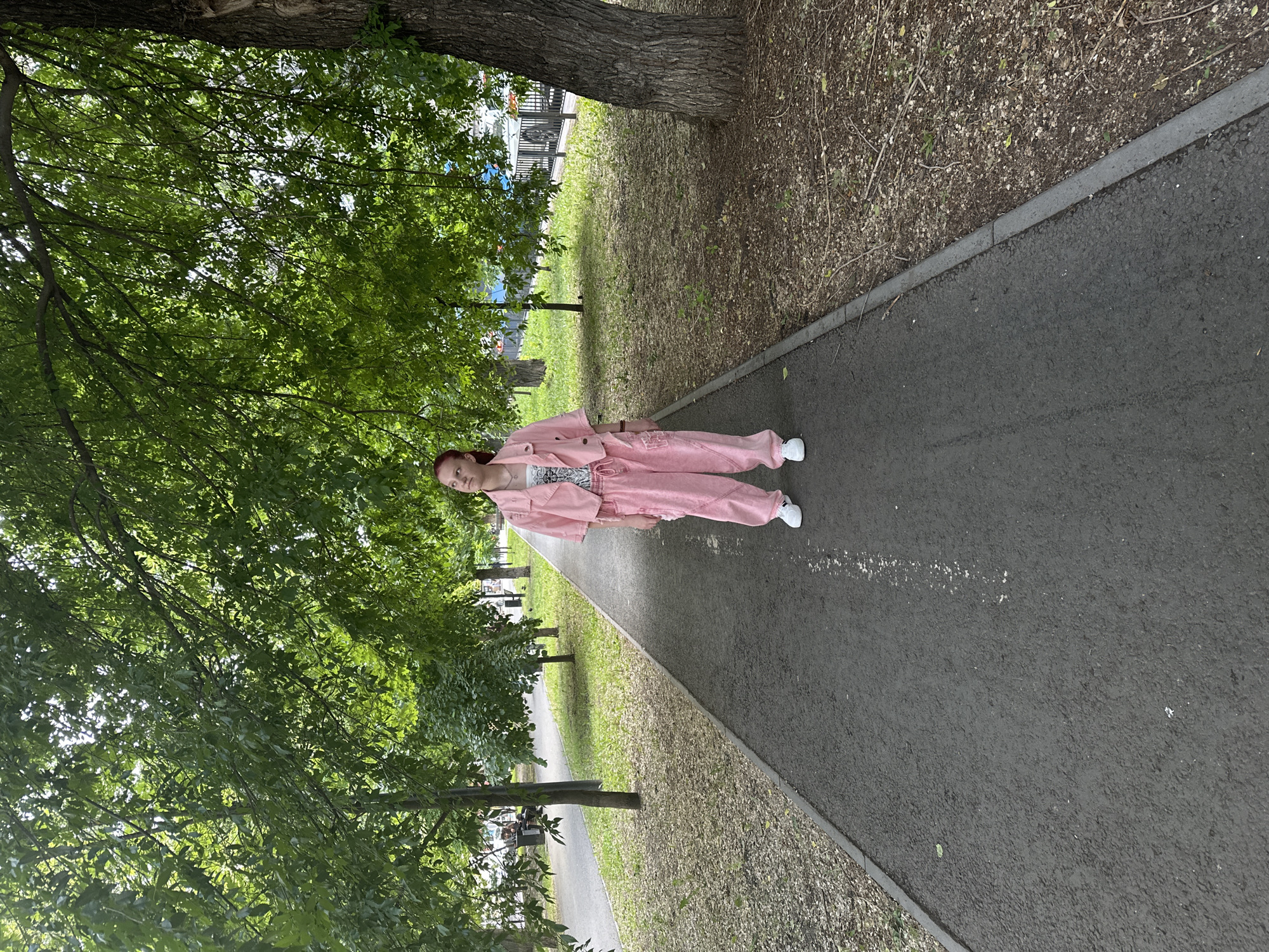 A person is standing on a paved pathway, likely a sidewalk, in a park-like setting. The individual is wearing a pink jacket and pants, with white sneakers. Green trees are abundant on both sides of the path, providing shade. The location is indicated as Saratov, Russia. The person is the primary subject. The environment consists of trees, grass, and a paved walkway.