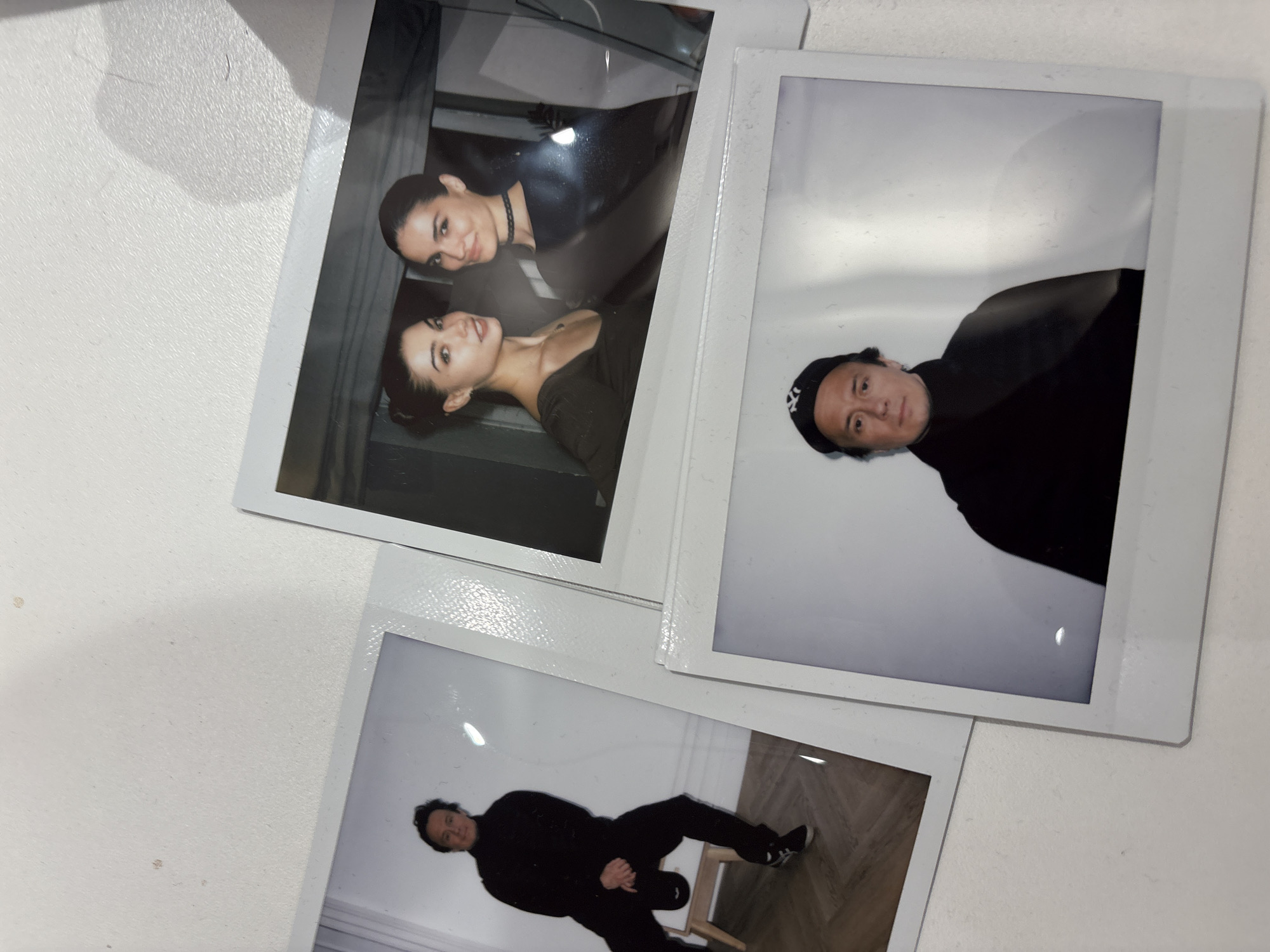 The image shows three Polaroid photos on a white surface. The first photo features two women in black tops, one wearing a choker. The second photo shows a man in a black turtleneck and a baseball cap. The third photo depicts a man in a black outfit sitting on a chair. The context of these photos in relation to Moscow, Russia is not available in the image.