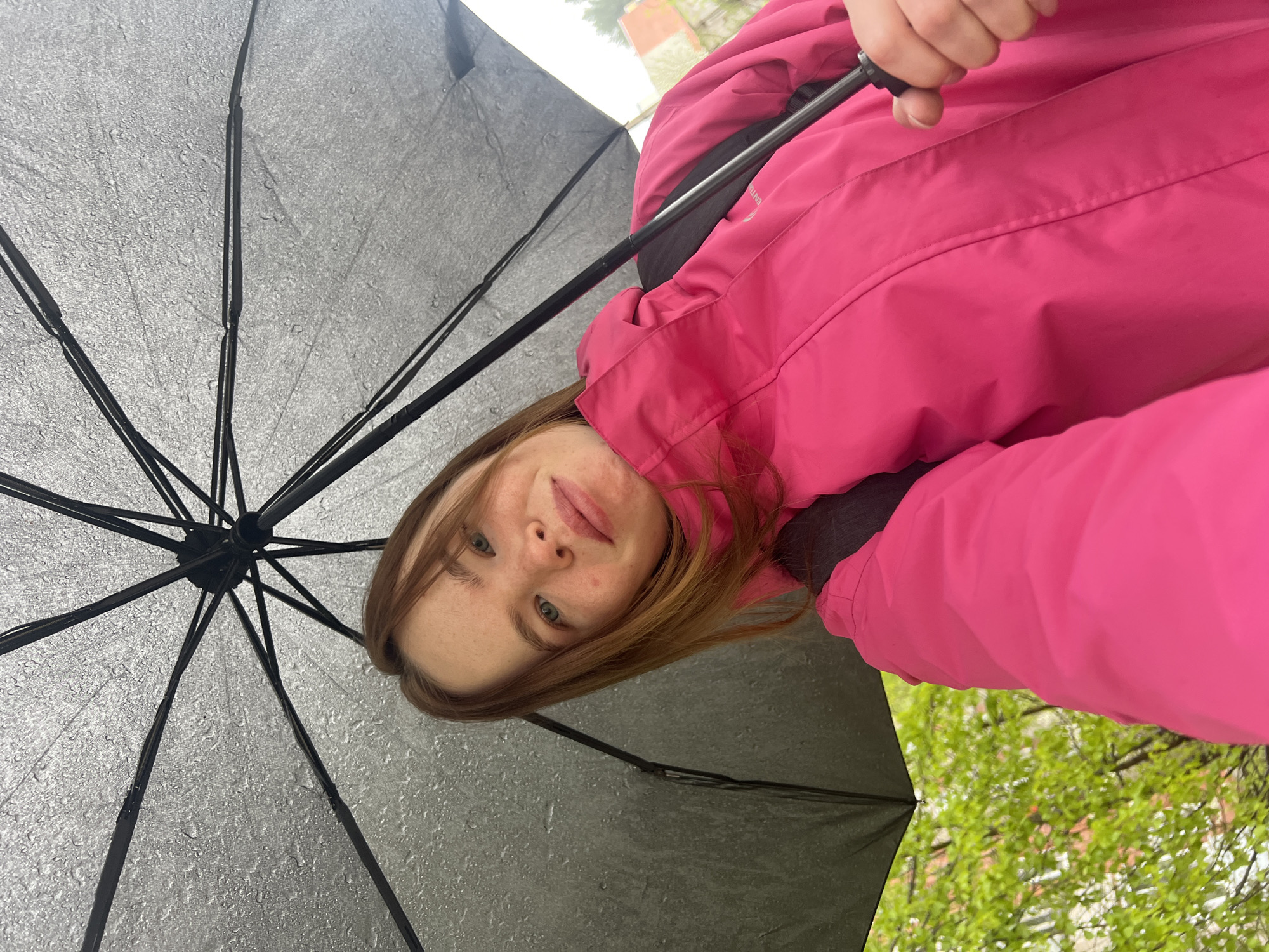 A woman with fair skin, freckles, and light brown hair is holding a black umbrella. The umbrella has raindrops on it. She is wearing a bright pink jacket. The background shows green trees and buildings, suggesting an outdoor location in the city of Chaikovsky, Russia. The image is taken from a low angle, looking up at the woman and the umbrella.