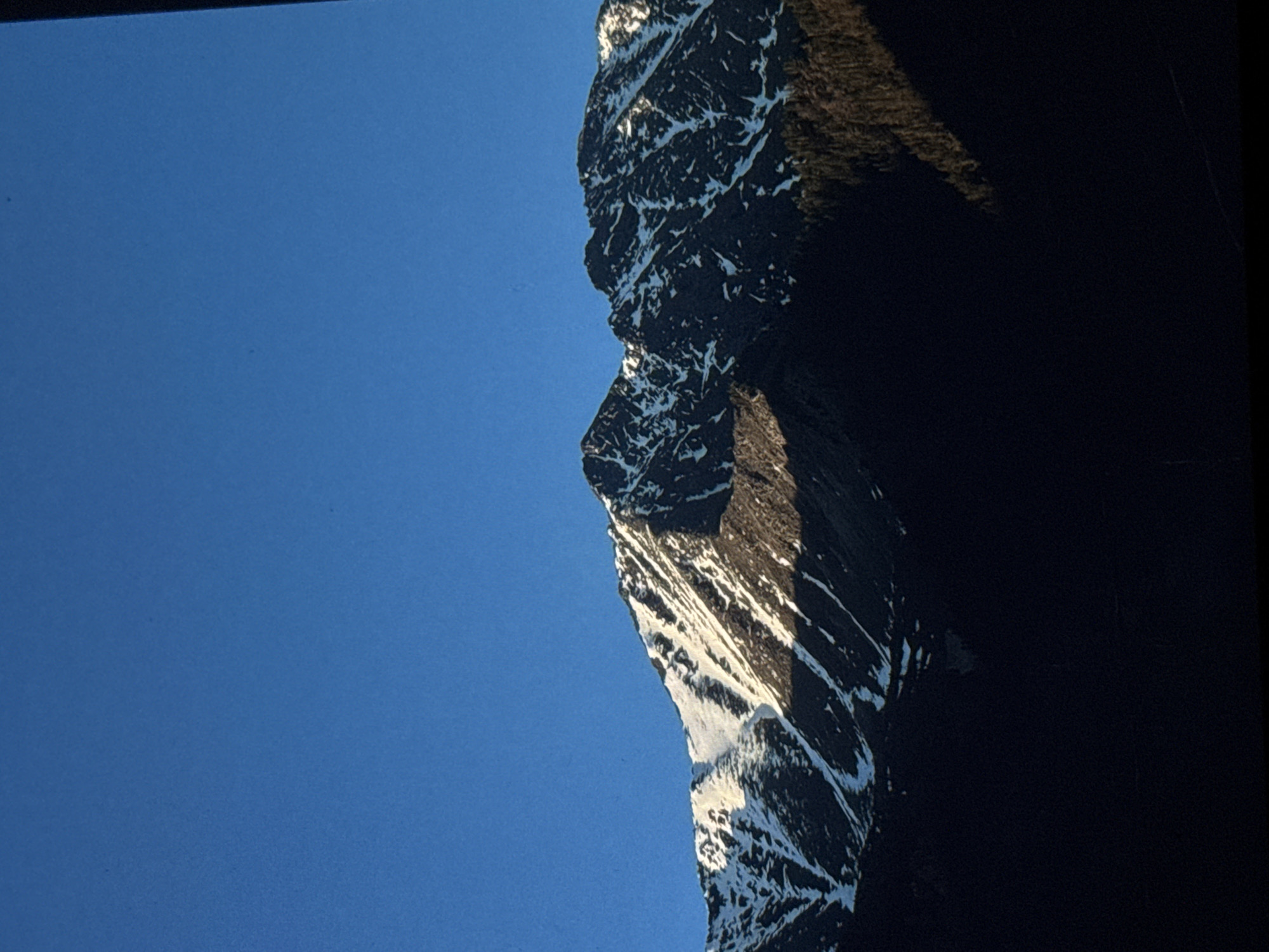 This is a vertical, close-up, outdoor, daytime photograph of snow-capped mountains under a clear blue sky. The mountains occupy the lower half of the image, with the sky taking up the upper half. The sunlight is hitting the upper slopes of the mountains, creating a contrast between illuminated and shadowed areas. The snow appears patchy and is present in streaks and patches along the rugged mountain faces. In the foreground, the lower slopes of the mountains are in deep shadow, obscuring any details of vegetation or terrain. The image does not depict any people, actions, or interactions. The location is not within the city of Irkutsk, Russia.