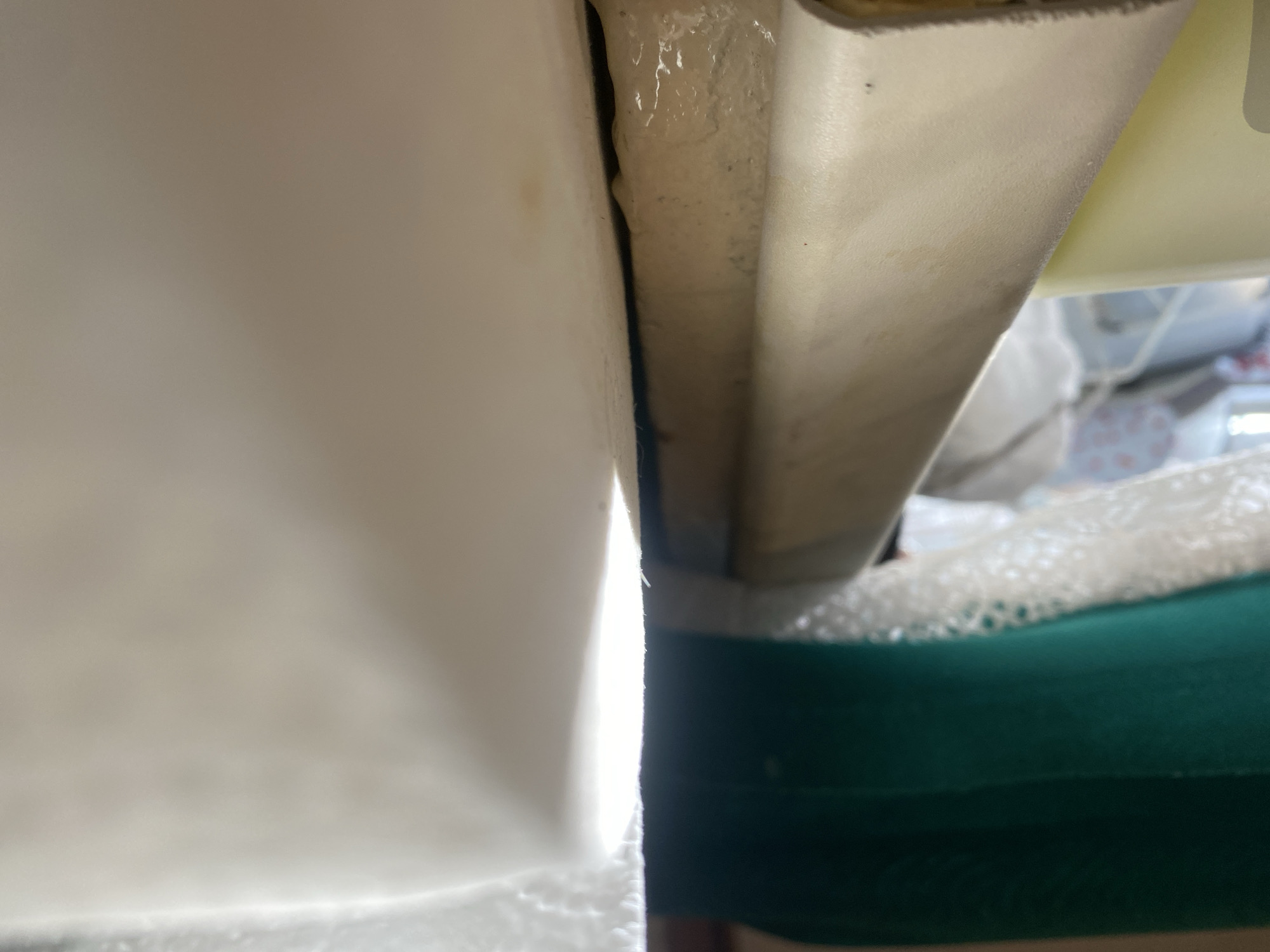 A close-up, low-angle shot focuses on a light-colored, textured surface that appears to be a rolled-up fabric or material. The surface is curved and shows some discoloration, with what looks like a sticky, translucent substance dripping from its edge. In the background, a blurred green fabric is visible, and behind that, other out-of-focus elements suggest an indoor setting. The lighting creates a strong highlight on the foreground surface, with a shadow cast to the right. The scene is static and does not depict any people or interactions.
