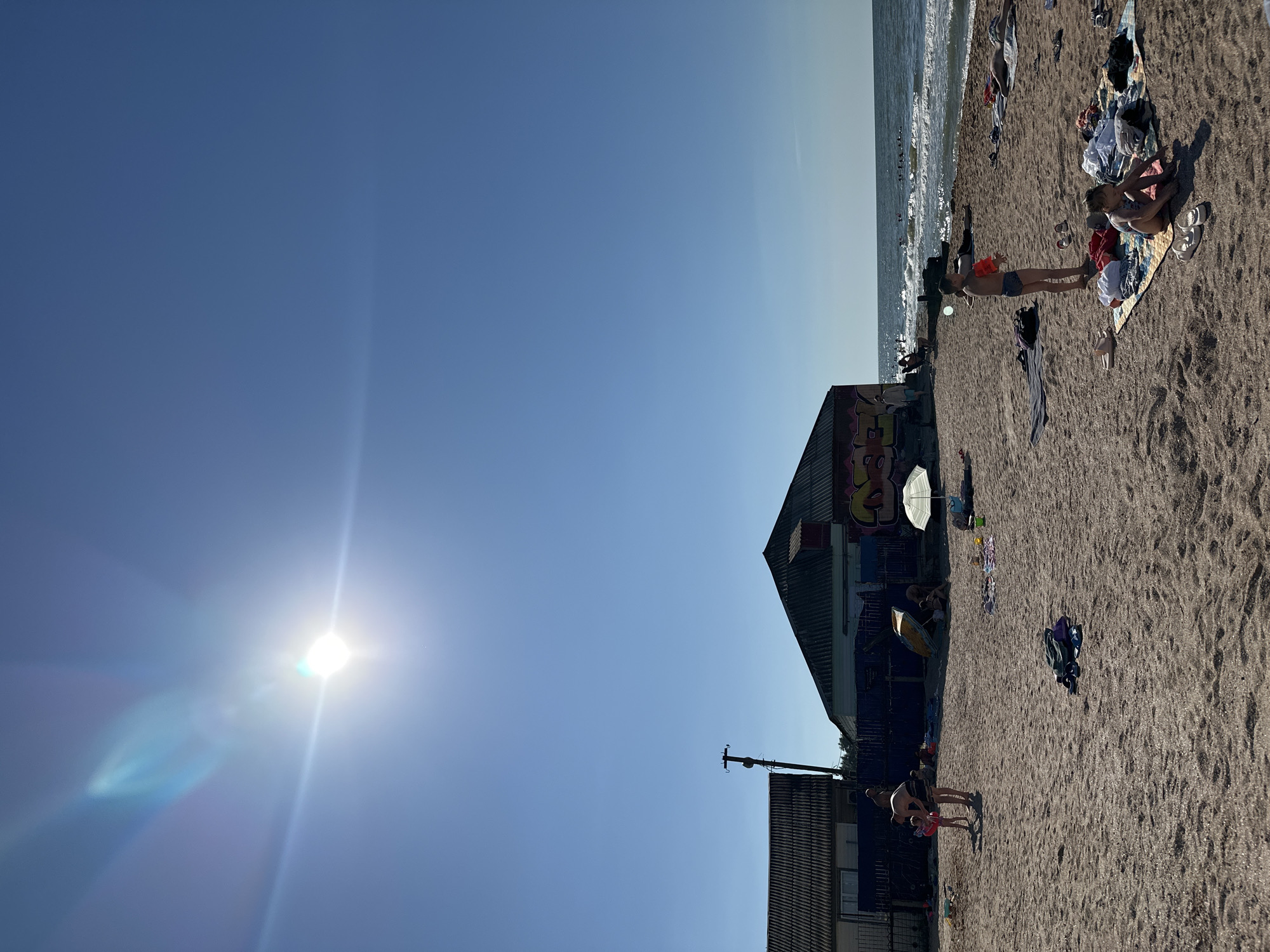 A sunny day at the beach in Golubitskaya, Russia, with the Black Sea in the background. The sand is covered with various beachgoers and their belongings. A building with graffiti is on the left side of the beach, and another structure is to its right. Several people are visible on the beach, including children playing and adults sunbathing. The ocean is calm, with gentle waves lapping the shore. The sky is clear and blue, with the sun shining brightly. The overall scene depicts a typical summer day at the seaside, with people enjoying leisure activities.