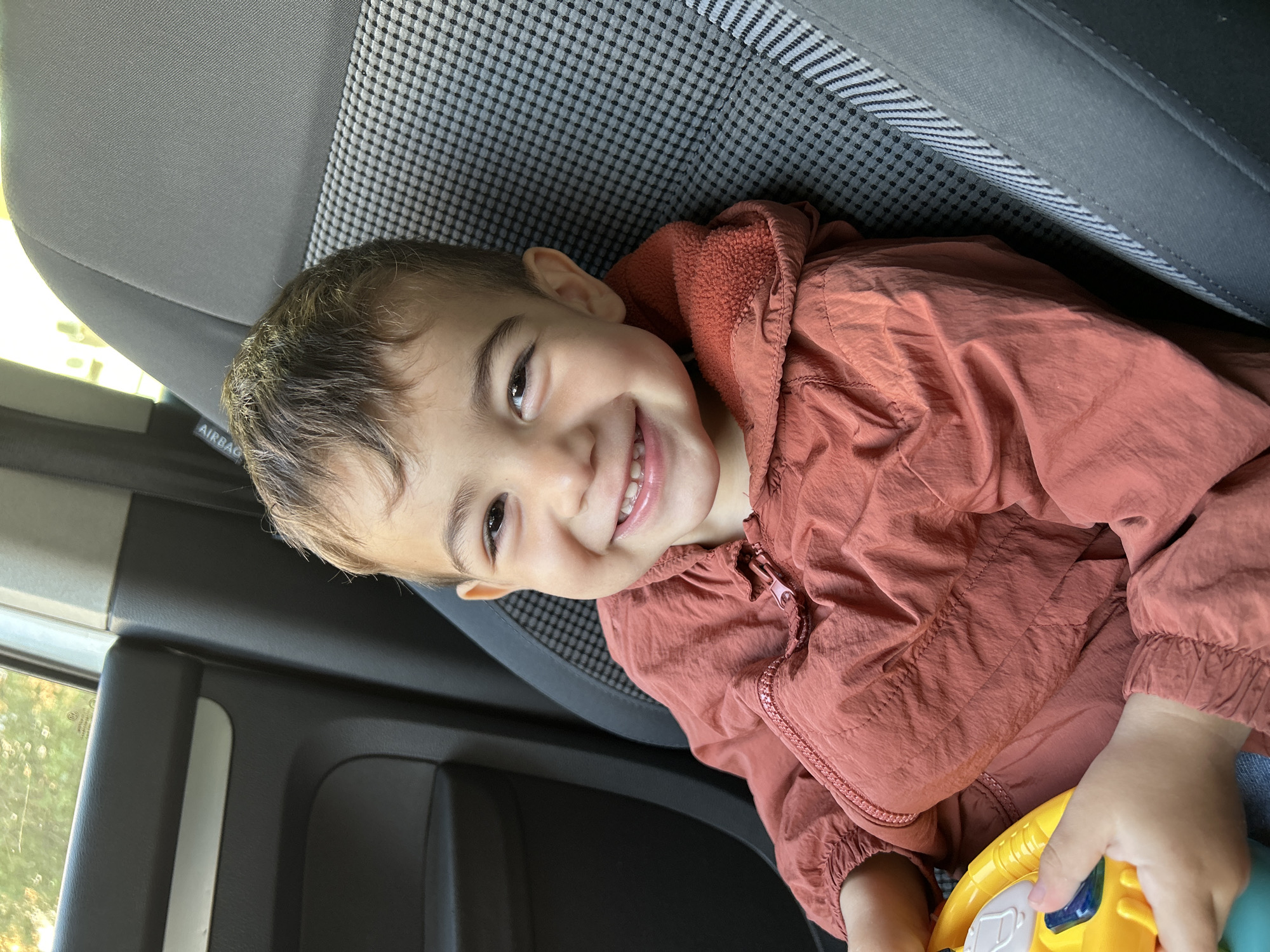 A young child, likely a toddler or preschooler, with light brown hair and dark eyes, is captured in a close-up shot inside a car, in Kardzhali, Bulgaria. The child is smiling broadly, showing their teeth, and looking directly towards the camera with a joyful and playful expression. They are wearing a brick-red or rust-colored hooded jacket made of a lightweight, crinkly fabric. Their hands are visible at the bottom right, holding a brightly colored toy, predominantly yellow and blue with some white and green details.
The child is positioned against a car seat, which features a dual-texture design: a plain grey fabric and a section with a distinctive black and white small checkered pattern. To the left, a car window offers a glimpse of bright daylight and out-of-focus green foliage, indicating an outdoor daytime setting. Parts of the dark grey car interior, including a door panel and seat structure, are also visible. The word "AIRBAG" is discernible on the side of the car seat, indicating a safety feature. The overall impression is one of candid childhood happiness within a vehicle.