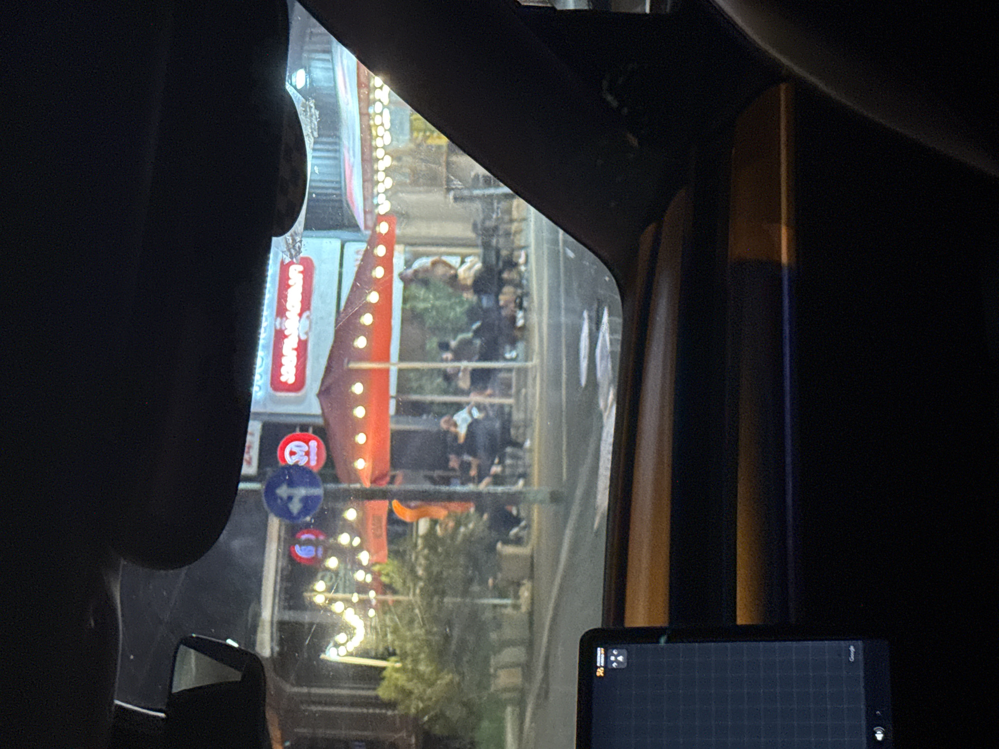 The image, captured from within a vehicle at night, shows an outdoor seating area of a restaurant or cafe in Tbilisi, Georgia. The foreground features a portion of a car's interior, including a dark dashboard and a digital display screen on the left. Beyond the vehicle's windshield, illuminated string lights stretch across the scene, casting a warm glow. A red umbrella, likely covering an outdoor dining space, is prominently visible. Several individuals are seated at tables, engaged in conversation or dining, though their features are indistinct due to the low light conditions and distance. Street signs, including a blue circular sign with a white arrow, are present. The background suggests a street or pedestrian area with buildings and more lighting. The overall ambiance is that of a lively evening gathering at an urban establishment.