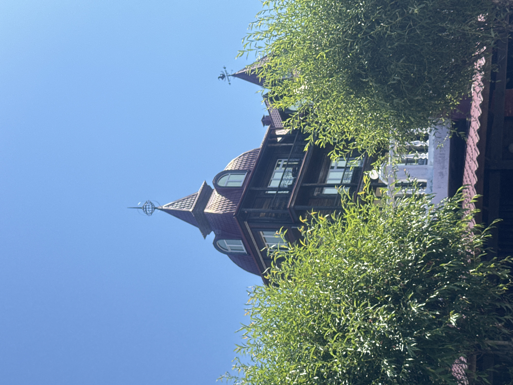 The image shows a wooden building with red roofs, reminiscent of a traditional Russian dacha, under a clear blue sky. The building has multiple stories with arched windows and balconies with ornate railings. Two prominent turret-like structures with pointed roofs top the building, one adorned with a decorative globe-like finial and the other with a weather vane shaped like a rooster. Lush green foliage from trees in the foreground partially obscures the lower portion of the building, including a white balustrade on a balcony. The scene is likely a sunny day, given the bright lighting and clear sky. While the specific location within Listvyanka, Russia, is not explicitly identifiable from the image alone, the architectural style suggests it could be a hotel, guesthouse, or a private residence in the scenic region. No people or actions are visible in the frame.