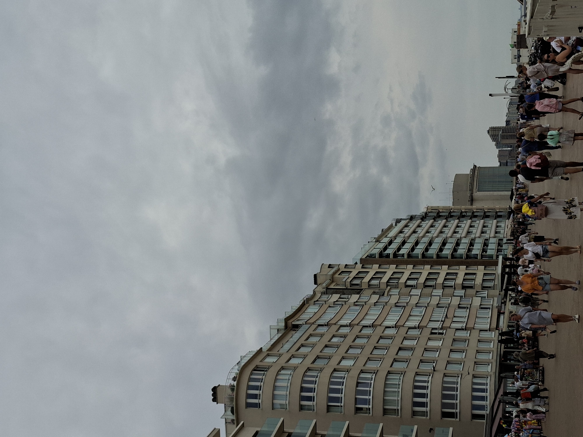 This is a ground-level, vertical shot of the Ostend, Belgium waterfront promenade under a cloudy sky. The promenade is lined with multi-story buildings, some with curved facades and glass balconies. The buildings are a mix of beige and white, with many windows. A flock of birds is visible in the sky. A large crowd of people is walking along the promenade, some in casual summer attire. There are market stalls on the left side of the promenade. In the distance, more buildings and structures are visible along the coastline. The overall atmosphere suggests a busy day at the seaside despite the overcast weather.