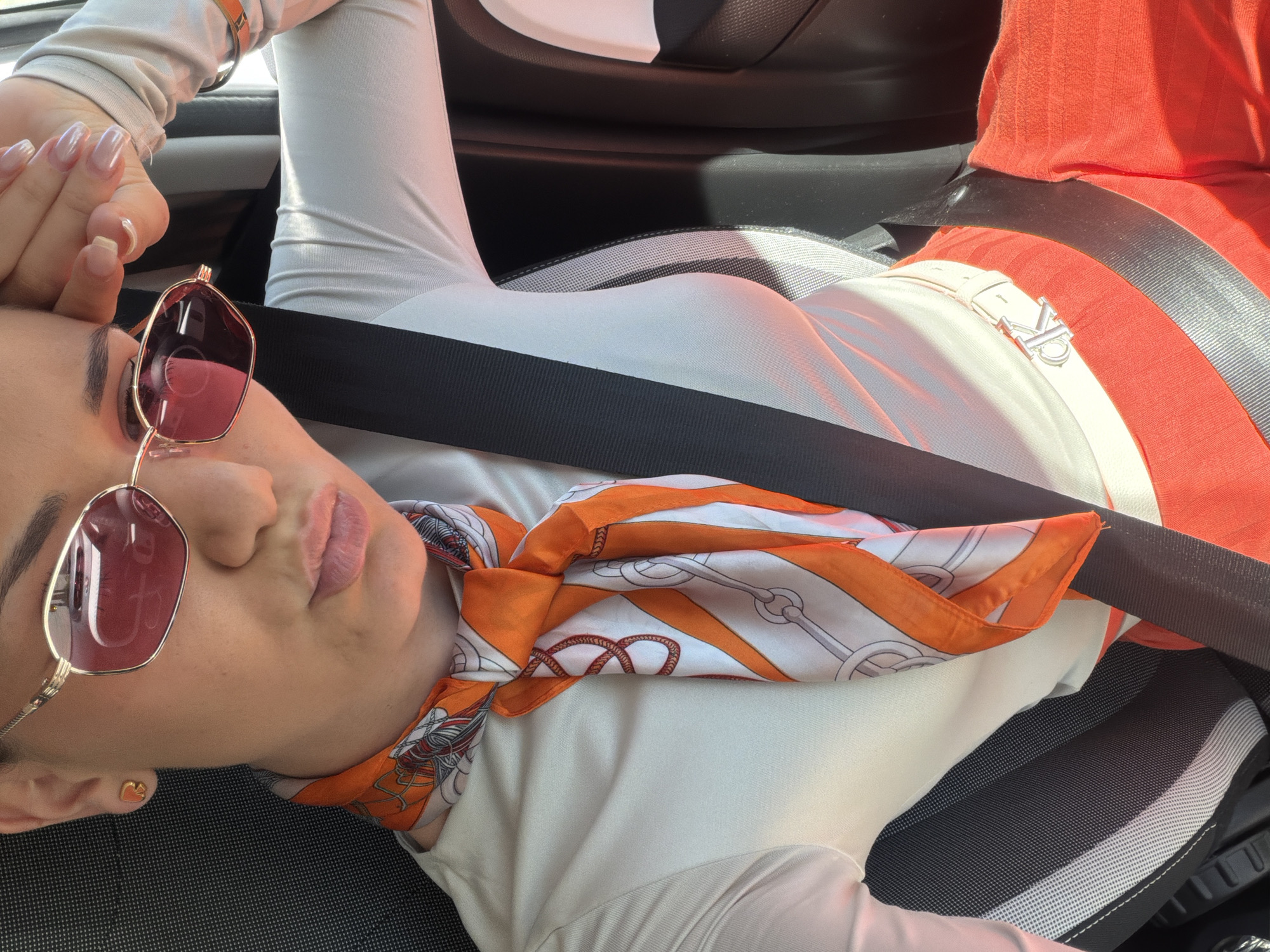 A woman is reclined in a car seat, wearing a white long-sleeved shirt, an orange and white patterned scarf, and rose-tinted sunglasses. She has her hand near her forehead and a purse expression on her lips. She is secured by a black seatbelt. The interior of the car is visible, including the car seat and door panel. There is no information to suggest that the image was taken in Sofia, Bulgaria.