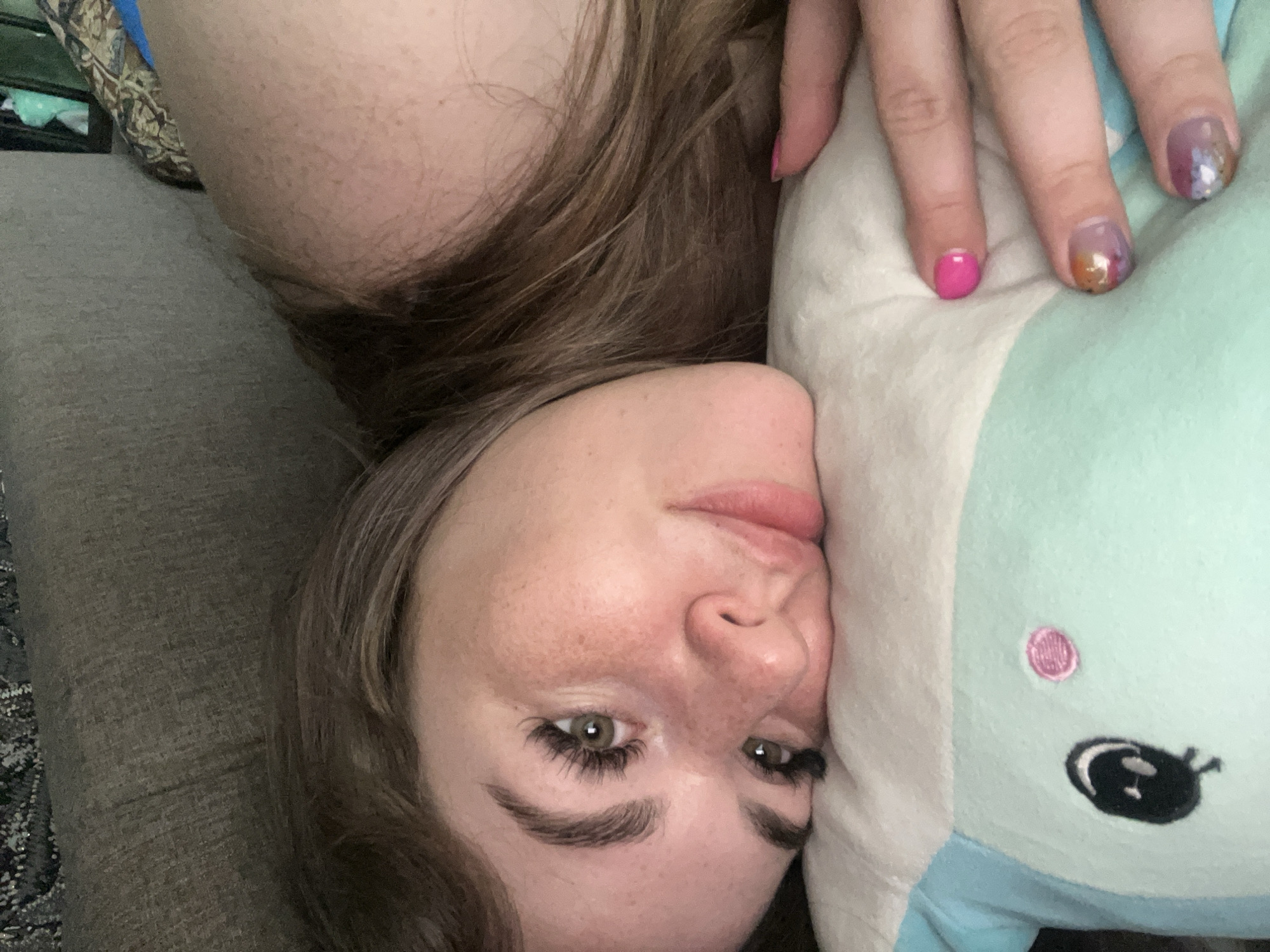 The media file presents an overhead, close-up portrait of a young woman lying on her side indoors, potentially in Chaikovsky, Russia. Her face, positioned horizontally in the frame, features green/hazel eyes, full lips, prominent eyebrows, eyelash extensions, and freckles scattered across her nose and cheeks. Her long brown hair is visible around her head and shoulders.
She is resting her head on a light blue plush toy, which has an embroidered black eye and a pink circular cheek. Her right hand is visible, draped over the plush toy, showcasing her fingernails. The pinky finger is painted a vibrant hot pink, while the ring and middle fingers display a distinctive shimmery, multi-colored design incorporating purple, orange, and gold hues.
The background consists of a light gray, textured sofa or bed. A patterned pillow or blanket in brown, gold, and blue tones is partially visible behind her head, and a dark, patterned rug or carpet can be seen in the bottom left. The lighting is soft and ambient, characteristic of an indoor setting. The woman exhibits a relaxed, neutral, or slightly pensive expression as she looks towards the camera. There is no visible text in the image.
