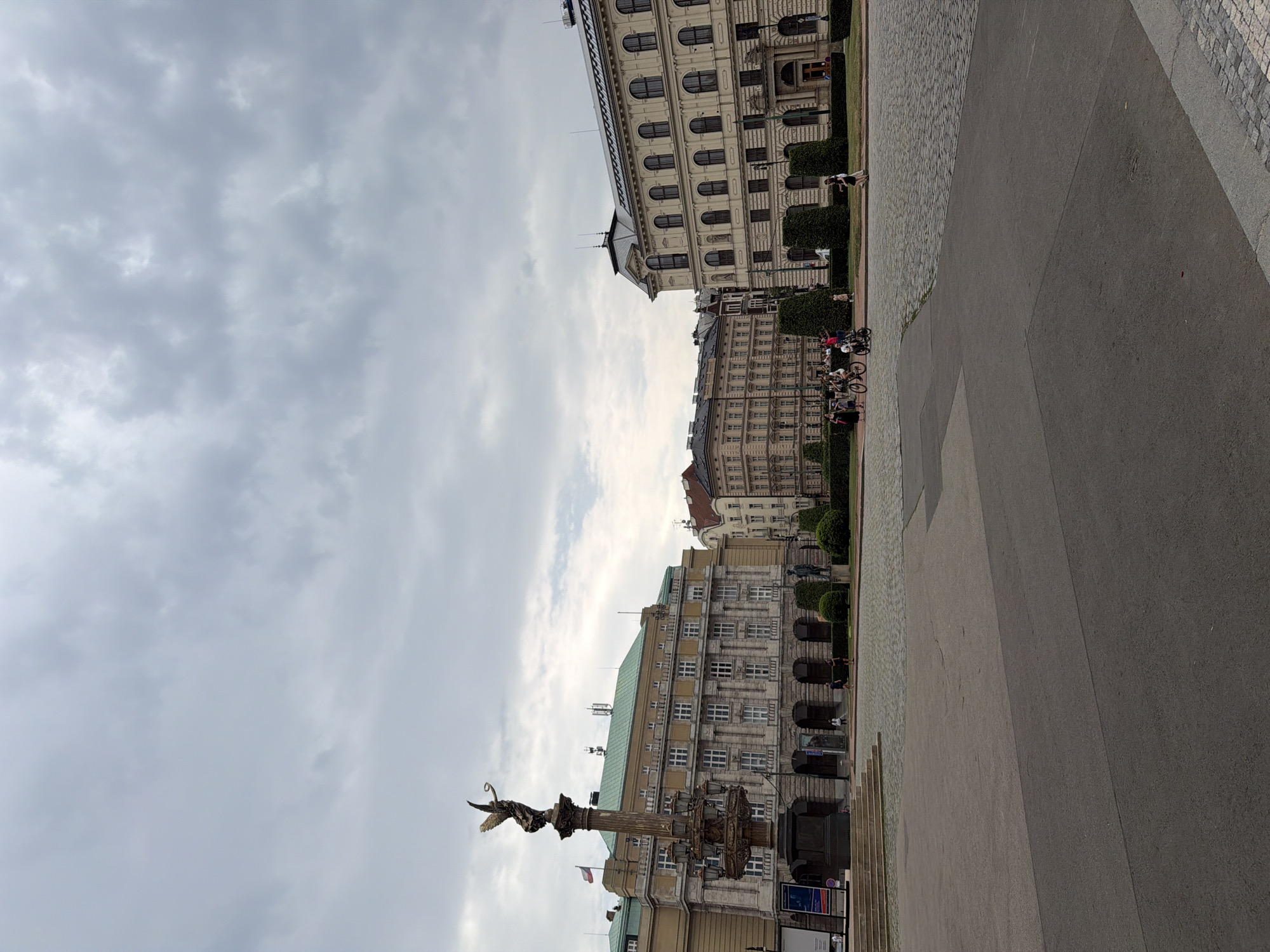 In Prague, Czechia, this photograph captures a wide shot of a city square under an overcast sky. In the foreground, a large paved area with asphalt gives way to a cobblestone section where several people are present. A group of individuals are walking and cycling on the cobblestones, with two people on bicycles and a few others walking nearby. To the left, a tall, ornate monument with a statue of an angel on top stands prominently, adorned with lamps and intricate carvings, and features a Czech flag on a flagpole. Behind the monument, a large, multi-story building with a light-colored facade, arched entrances at the ground level, and numerous windows dominates the middle ground. Further to the right, another similar, large building with multiple stories and many windows is visible, featuring an arched entryway. The background consists of more buildings and a dense canopy of clouds. The scene is an urban environment with architectural structures and public space.