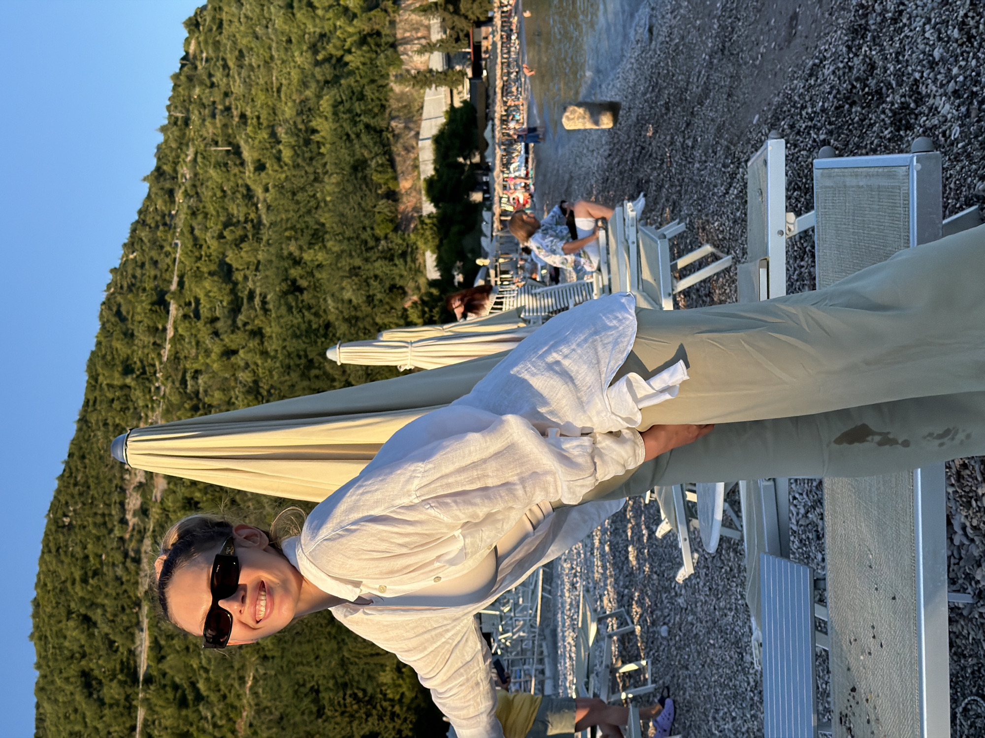 A woman in sunglasses and a white open-collared shirt over a light-colored top and olive-green pants is leaning forward on a pebble beach in Budva, Montenegro. She is smiling and looking at the camera. Behind her, rows of unoccupied lounge chairs with umbrellas are set up. In the background, a crowded beach with people swimming and sunbathing is visible, with a densely forested hillside rising steeply behind it. The sky is clear and blue, indicating it is daytime.
