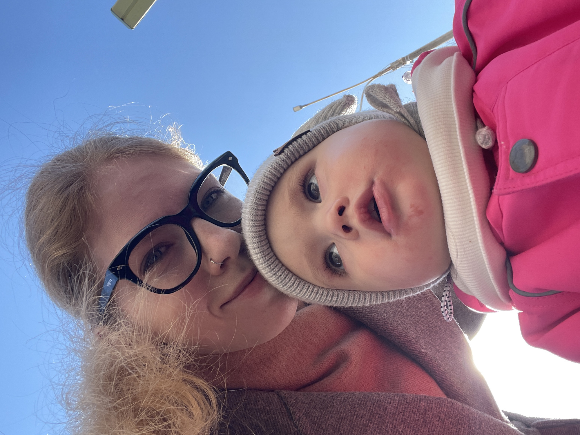 A close-up, upward-angle photograph shows a woman and a young child. The woman, positioned on the left, wears black-framed glasses and a nose ring. She has blonde hair and is wearing a dark colored sweater. The child, possibly being held by the woman, wears a grey knit cap with bunny ears and a bright pink coat with white trim. The sky is visible in the background, suggesting an outdoor setting.
There is no indication in the photo that would suggest the location is Izhevsk, Russia.