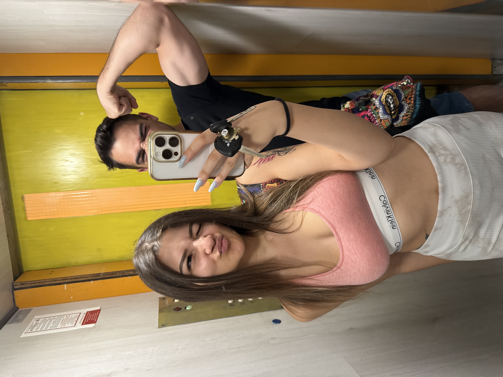 In an elevator in Sofia, Bulgaria, a woman with long brown hair and a pink sports bra winks at the camera. She is holding a smartphone and a set of keys. Behind her, a man in a black t-shirt flexes his bicep, his face partially obscured by the phone. The elevator walls are a vibrant yellow and orange, contrasting with the grey paneling.