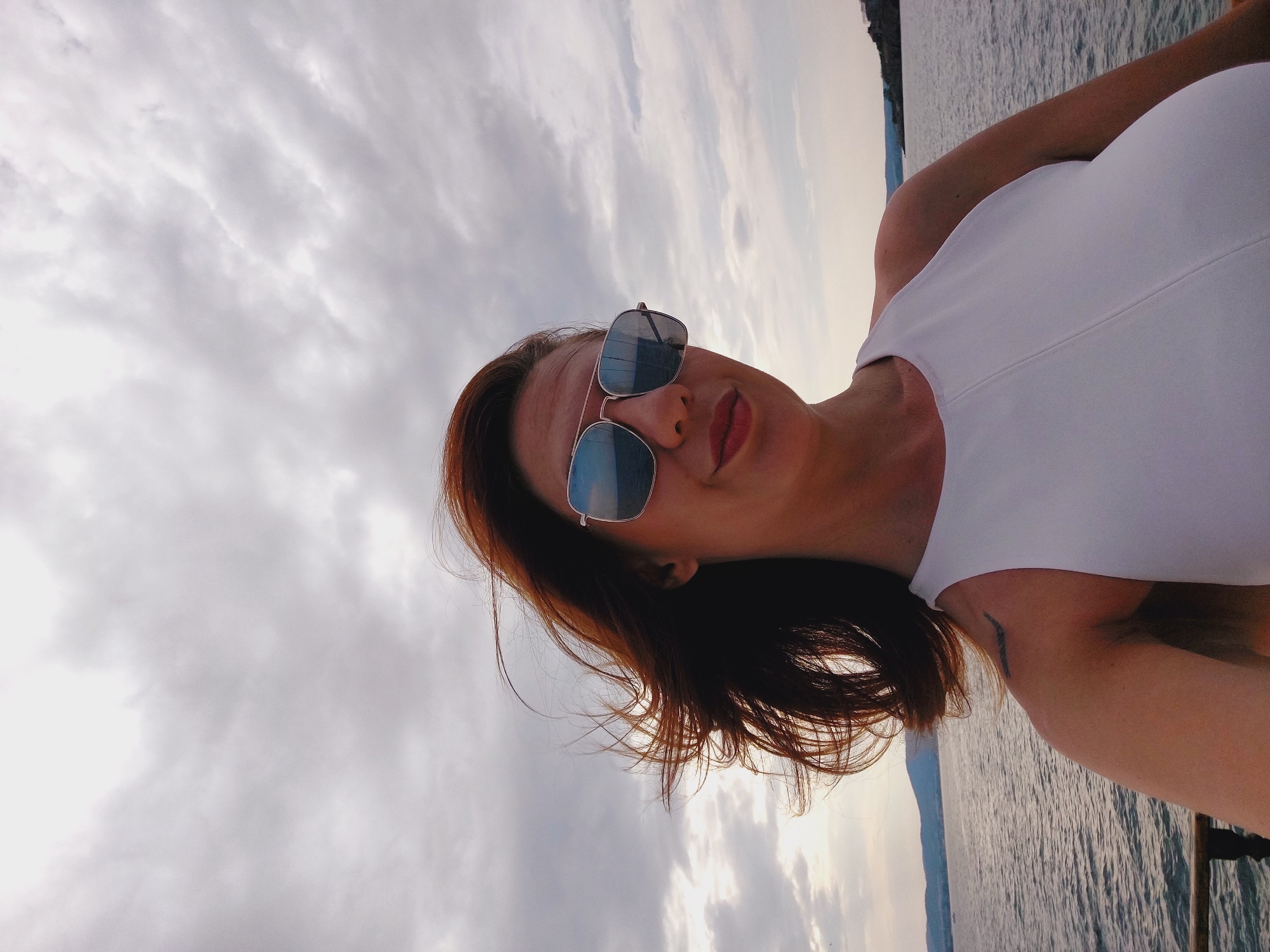 A woman with red hair and sunglasses is taking a selfie outdoors. She is wearing a white top and smiling. The background shows a body of water and a cloudy sky. The location is likely the coast of Sozopol, Bulgaria, given the presence of the sea and the style of the landscape visible.