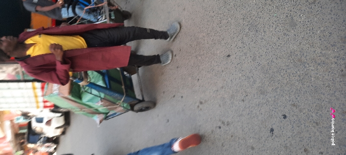 The image captures an urban street scene in Nairobi, Kenya, depicting pedestrians and a loaded handcart. The central figure is a dark-skinned male, visible from the mid-thigh up, wearing a dark beanie, a yellow short-sleeved polo shirt, a long maroon open coat, dark pants, and light grey slip-on shoes. He is walking towards the right side of the frame, with his right arm bent. To his left is a blue metal-framed, two-wheeled handcart, heavily loaded with green tarp-covered bundles secured by rope or cord. In the lower-left foreground, the lower section of a person's left leg is visible, clad in blue jeans and a shoe with a prominent reddish-orange sole and white upper. The background is blurred, showing a white sedan, a truck with vertical white and yellow stripes, and faint outlines of structures consistent with urban buildings or market stalls. A partially visible individual is present on the far right, appearing to be on a two-wheeled vehicle or another cart. The ground consists of a grey paved surface with various minor stains and textures. The text "police kombo💖💖" is present in white in the lower-left corner.