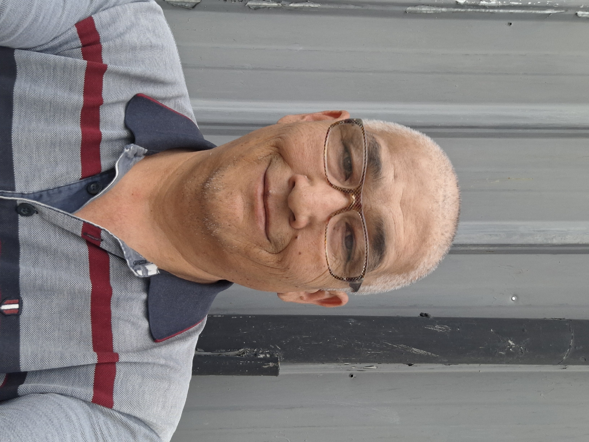 A middle-aged East Asian man wearing glasses and a collared polo shirt poses for a selfie in front of a corrugated metal wall. The man has short, graying hair and a light stubble. The polo shirt is gray with horizontal dark red and navy blue stripes. He is smiling. The background is a neutral gray metal wall, with a black vertical pipe or beam on the right side. The lighting appears to be overcast. The image was taken outdoors in Almaty, Kazakhstan.