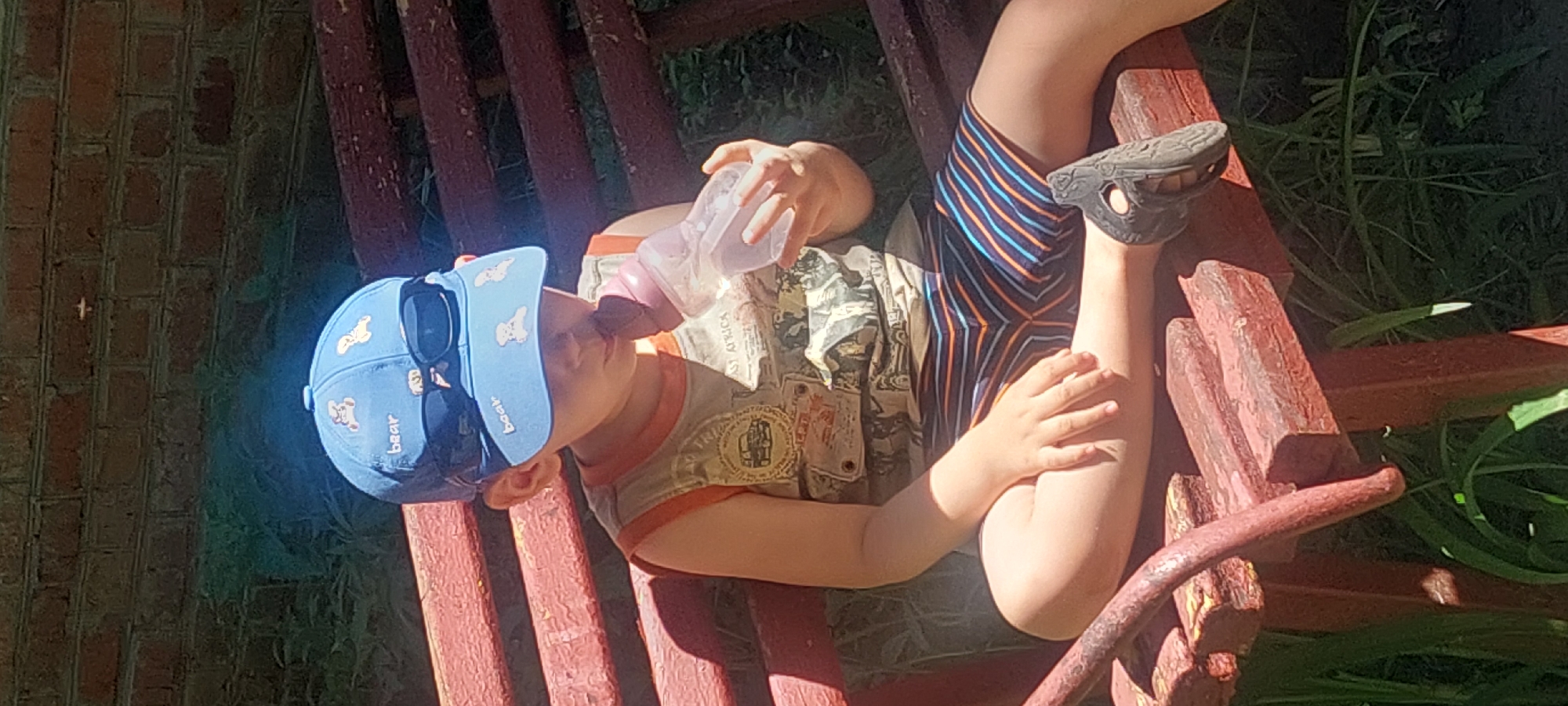 The image displays a young child seated outdoors on a weathered reddish-brown wooden bench. The child is wearing a blue baseball cap featuring white cartoon bear figures and the word "baar" embroidered in white. Dark-lensed sunglasses are positioned on the brim of the cap. The child wears a light-colored, sleeveless t-shirt with orange trim and graphics that include depictions of vintage vehicles and partially legible text such as "EAST AFRICA." Striped shorts, predominantly blue with orange and light blue lines, cover the child's lower body, and dark-colored sandals with perforations are on the child's feet. The child is holding a clear plastic baby bottle with a pink nipple to their mouth, appearing to drink from it. In the background, a red brick wall built in a stretcher bond pattern is visible, with green grass and foliage growing beneath it. A small, light-colored circular object is noticeable on the brick wall. The scene is illuminated by strong, direct sunlight, creating distinct shadows. No specific identifiers or landmarks are present in the image to determine a precise location within Kharkiv, Ukraine.