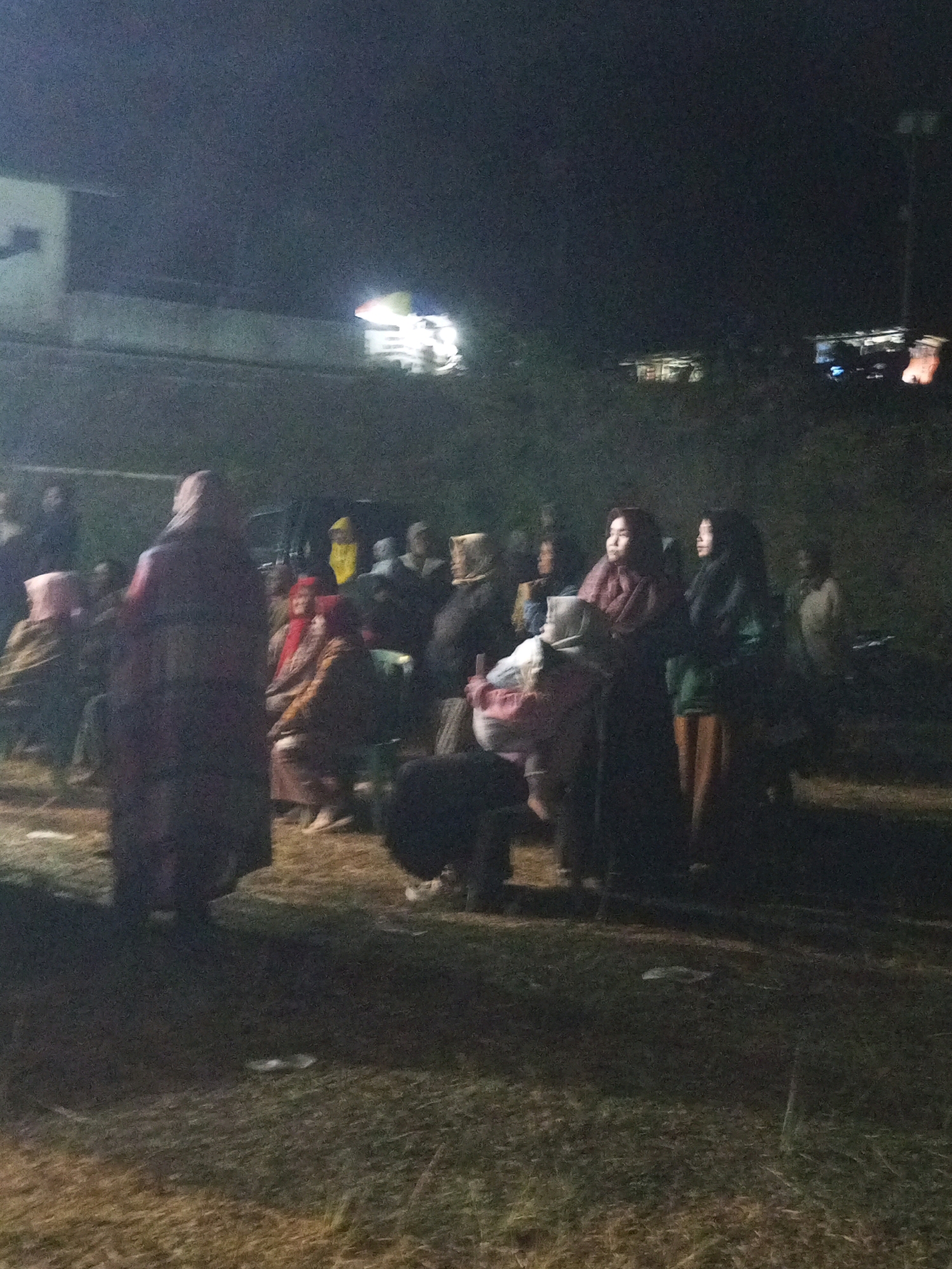 In Ngadikerso, Indonesia, a dimly lit nighttime scene captures a gathering of people, some seated and others standing, in an outdoor area. The atmosphere appears informal, possibly a community event or a casual social interaction. A faint light source, possibly from a lamp or building, illuminates the group from the left and rear. Several individuals are clad in traditional attire, with some women wearing headscarves. A structure, possibly a building, is visible in the background on the left, with lights suggesting occupancy. The ground appears to be uneven terrain covered with grass and scattered debris. The overall impression is that of a communal gathering taking place in a rural or semi-rural setting at night.