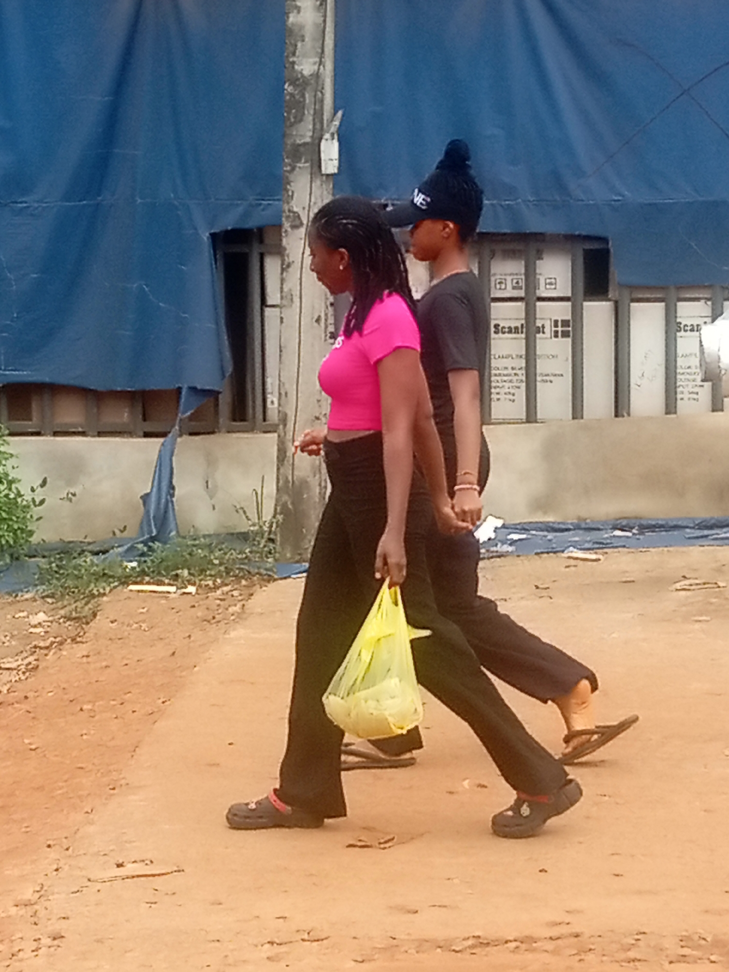 Two individuals are walking side-by-side on a dirt road in Umuokpu, Nigeria. The person on the left is wearing a pink shirt and black pants, carrying a yellow plastic bag. The person on the right is wearing a black shirt, black pants, and a dark-colored cap. Both are wearing dark-colored footwear. A blue tarp is draped over a building behind them. Boxes are stacked against the wall of the building with the tarp on it. The ground is uneven and dusty.
