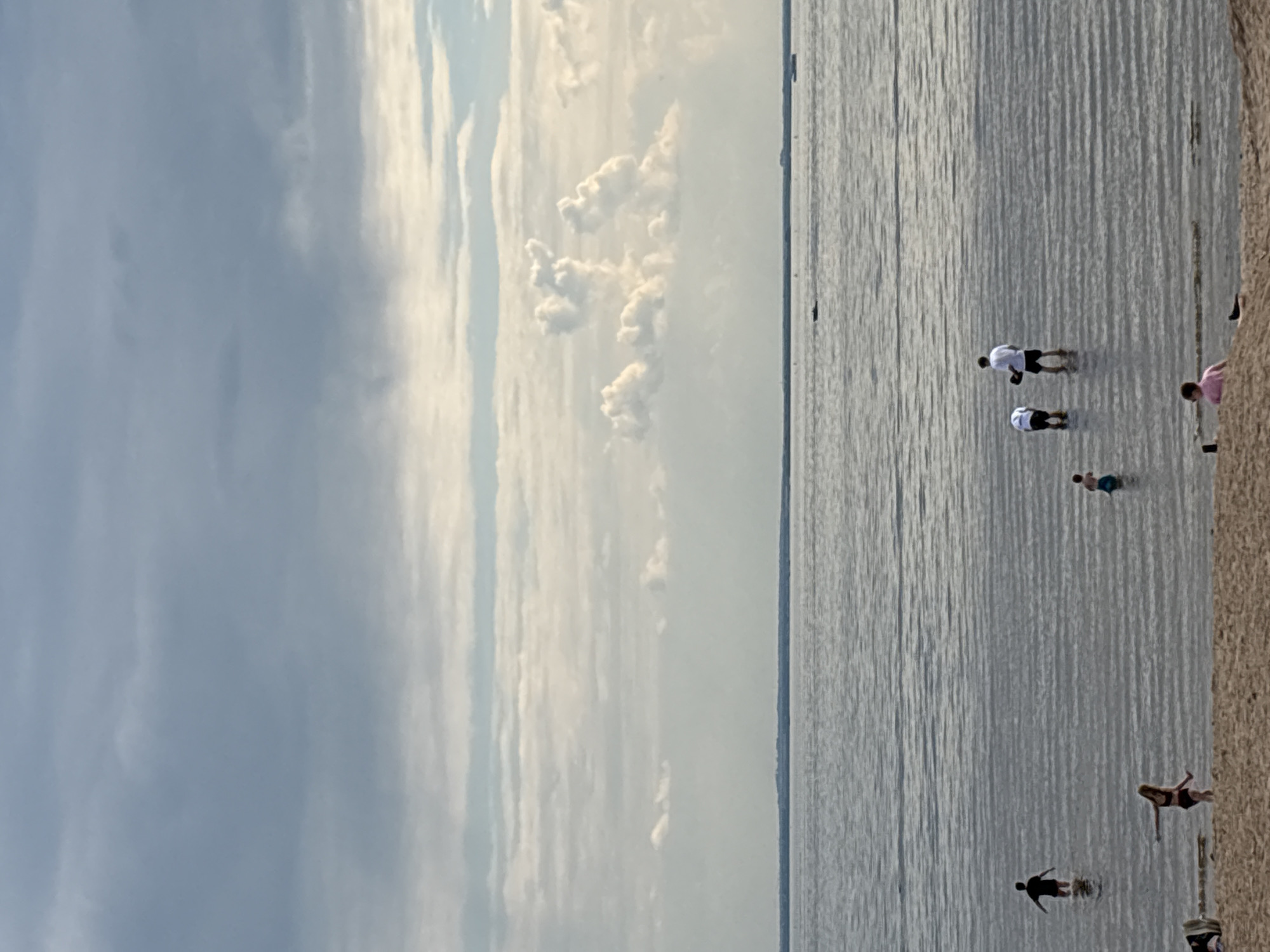 The image displays a serene beach scene, likely along the coast near Saint Petersburg, Russia, depicting a wide expanse of water meeting a sandy shoreline under a partly cloudy sky. The water, calm with subtle ripples, appears silvery-grey, reflecting the soft light from the sky. A distant horizon line separates the water from the sky, which features a mix of soft grey clouds and brighter, more defined white cloud formations.
Several people are enjoying the tranquil environment. In the shallow waters, two individuals, one in a white top and dark shorts and another appearing to be a child, are wading further from the shore. Closer to the sandy bank, another small child is partially submerged in the water. On the sandy edge, a person in a pink top is partially visible, while another individual in dark swimwear is seen sitting or lying near the water's edge. Towards the left foreground, a person in dark clothing is also wading into the water.
The lighting suggests late afternoon or early evening, characterized by a gentle, diffused illumination that contributes to a peaceful atmosphere. The weather appears overcast with breaks in the clouds allowing light to filter through. No distinct objects beyond the individuals and their attire are visible. The scene captures a quiet, leisure activity by the water. There is no visible text in the image.