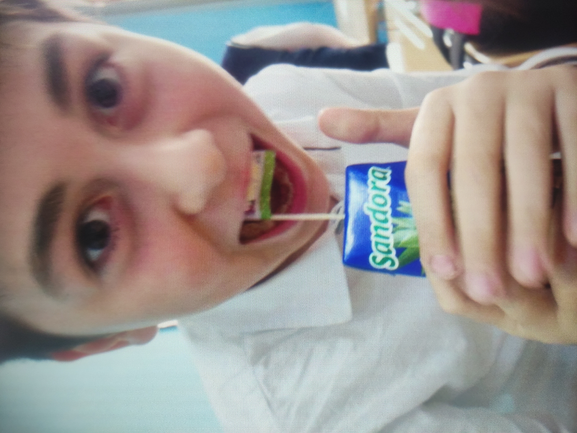 The image shows a close-up of a young boy holding a small juice box with the brand name "Sandora" visible. He is wearing a white collared shirt and has a piece of food, possibly a candy or a piece of fruit, in his mouth. His right hand is holding the juice box, and his left hand is visible supporting it. The background is blurred but appears to be indoors with a light blue wall on the right. There is no discernible information about the location in Baku, Azerbaijan.