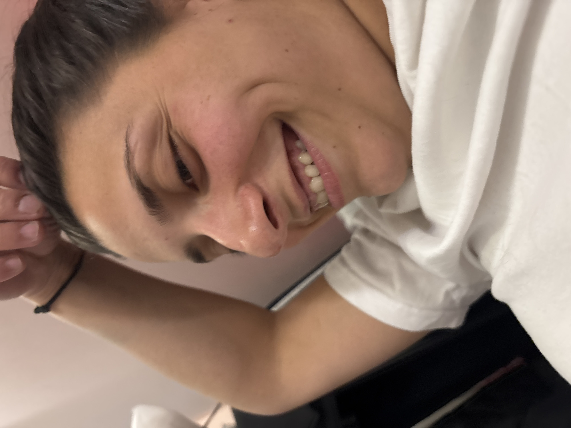 A medium close-up, high-angle shot depicts a smiling individual with dark hair and a fair complexion. The person is angled slightly towards the left, revealing their face and upper body. They are wearing a white t-shirt. The background is blurred, showing hints of a white surface or wall, a lamp, and dark clothing. A black bracelet is visible on their left wrist. The scene appears indoors, with ambient lighting. There is no indication of the city of Sofia, Bulgaria in this image.