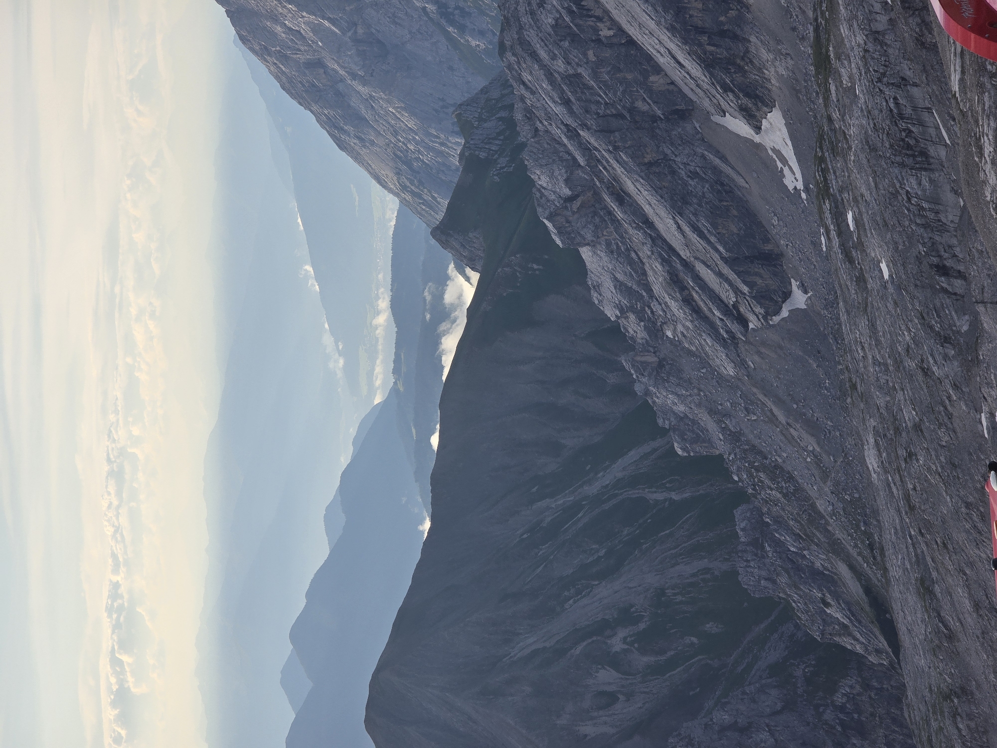 The image depicts a panoramic view of a mountainous landscape, likely taken from a high vantage point in or near Garmisch-Partenkirchen, Germany. The foreground features rugged, rocky terrain with patches of sparse vegetation and lingering snow. In the middle ground, steep, dark mountain slopes dominate, with deep ravines and visible geological stratification. Beyond these immediate peaks, layers of progressively lighter blue, mist-shrouded mountains recede into the distance, creating a sense of immense depth and scale. The sky above is a pale blue, with soft, diffused clouds, suggesting either early morning or late afternoon light. A portion of a red object, possibly a railing or part of a structure, is visible in the lower right corner of the frame, indicating the presence of a viewing platform or a facility for visitors. No people are clearly visible in the frame. The overall atmosphere is one of natural grandeur and serenity.