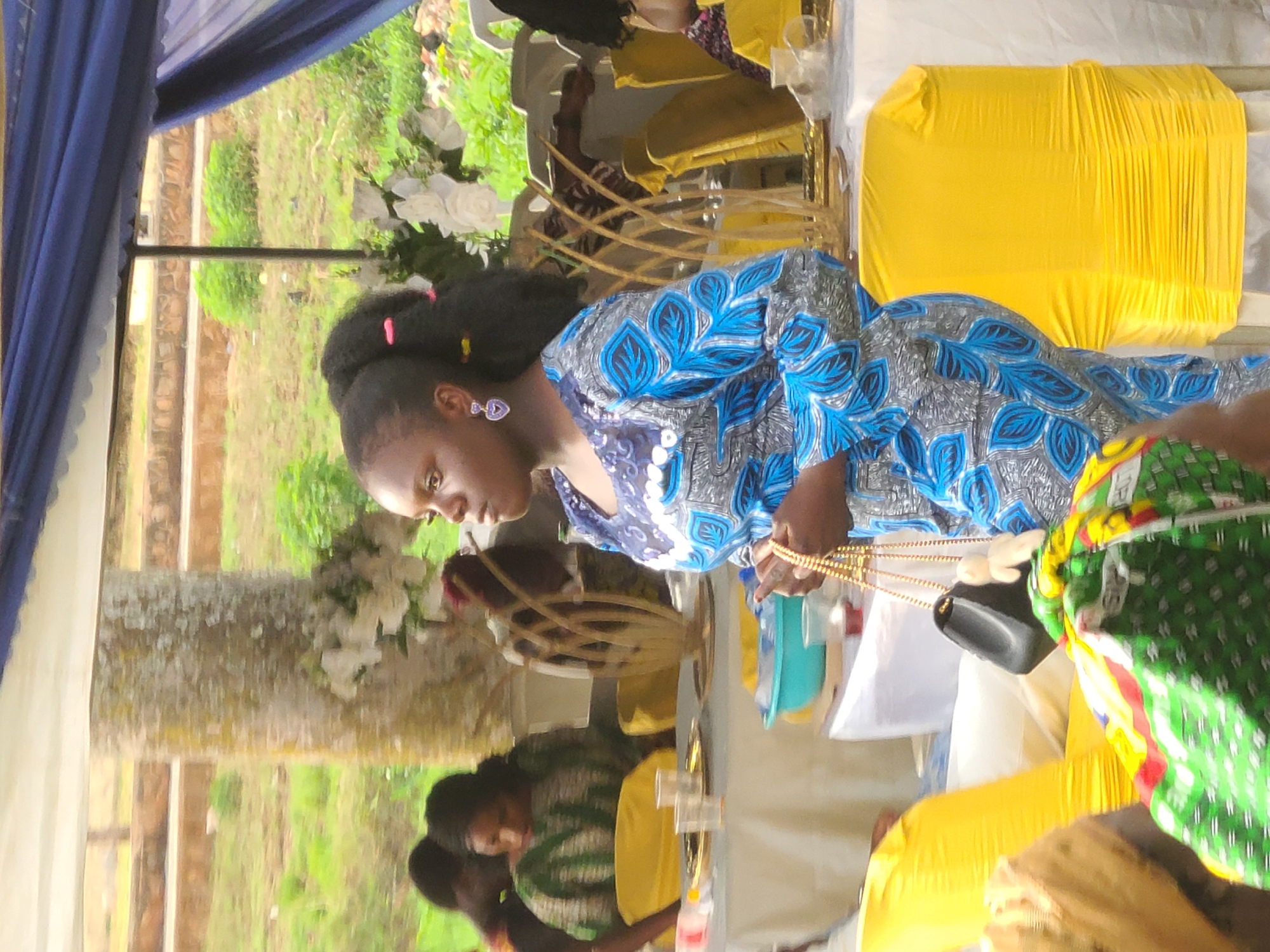 A woman with dark hair pulled back and wearing a blue patterned dress is seated at a table. She is holding a gold necklace and a small black bag. Other people are seated at the table, which is covered with a white cloth. Yellow chair covers are seen. The setting appears to be outdoors under a blue tent. A tree and steps are visible in the background. The location is likely in Jos, Nigeria, but the details are insufficient for confirmation.
