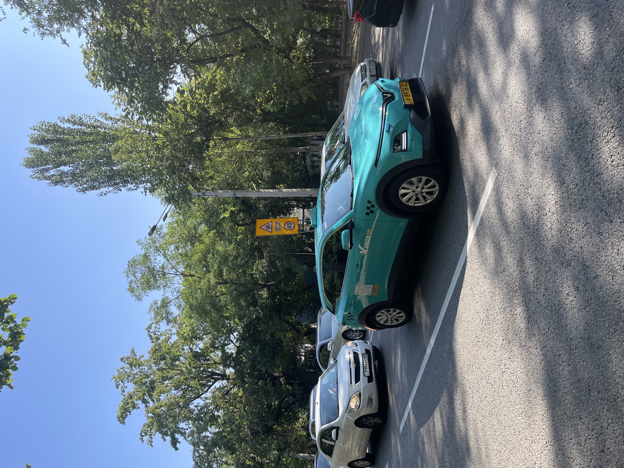 The image depicts a street scene in Almaty, Kazakhstan, under a clear blue sky. A bright turquoise electric taxi, identified by "GREENSM" branding and a QR code, is prominently featured in the foreground. This taxi has the license plate "P 8976 02". To its left, a white Chevrolet sedan with the license plate "719 AVT 17" is visible, along with other cars in traffic. In the background, lush green trees line the street, and a yellow traffic sign with the text "МЕКТЕП" and "40" is visible. The road surface is asphalt with white lane markings. The overall impression is a sunny day in an urban environment with active traffic.