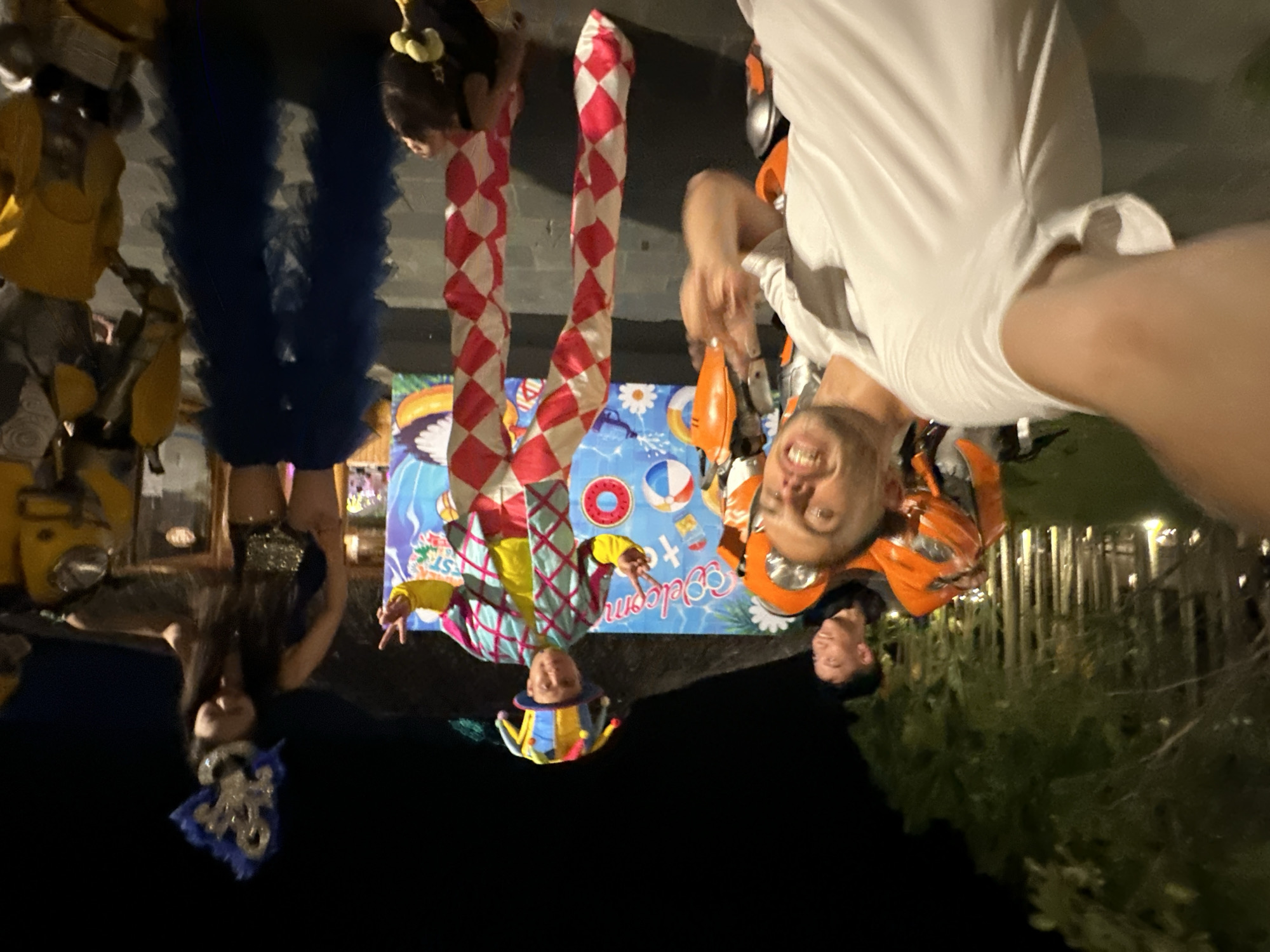 In Hoa Lư Ward, Vietnam, a nighttime outdoor event features a diverse group of performers and attendees. The foreground is dominated by a man in a white t-shirt, capturing a selfie with a wide grin and flashing a peace sign. Behind him, an individual dressed as a transformer, primarily in orange and black, is visible. Further back, a clown performer stands on stilts, wearing a colorful, checkered costume and a jester's hat. To the right, a woman in a dark outfit and an elaborate blue and silver headdress stands near another transformer costume, this one predominantly yellow. A backdrop behind the performers displays a "Welcome to Fest" banner with beach-themed imagery. The environment suggests a festive atmosphere with artificial lighting illuminating the scene, and trees forming a dark outline against the night sky.